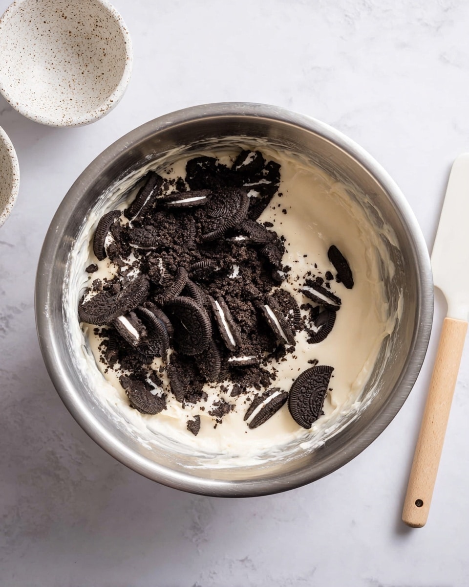 A large silver mixing bowl is filled halfway with smooth, creamy white batter, and on top, a layer of broken pieces of dark chocolate sandwich cookies with white cream filling is scattered unevenly. The bowl rests on a white marbled surface. A white kitchen spatula with a light wooden handle lies next to the bowl, and a speckled white small bowl is partially visible near the top left corner. The scene has soft, natural lighting. photo taken with an iphone --ar 4:5 --v 7