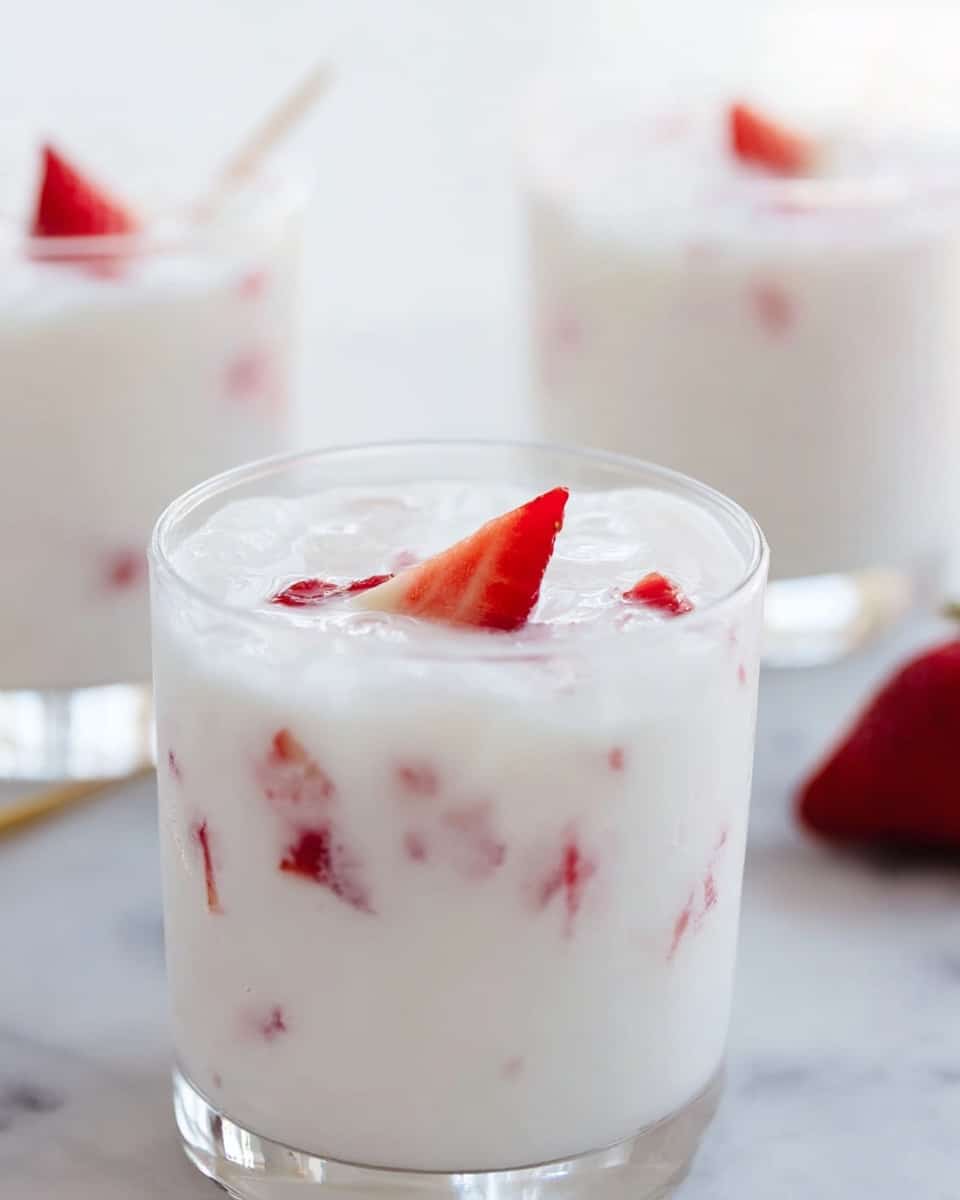 A clear glass filled with a white creamy drink with small pieces of red strawberry floating inside, topped with a small triangular slice of strawberry. In the background, there are two more similar glasses, all set on a white marbled surface. The light is soft and natural, highlighting the smooth texture of the creamy drink and the bright red of the strawberries, photo taken with an iphone --ar 4:5 --v 7