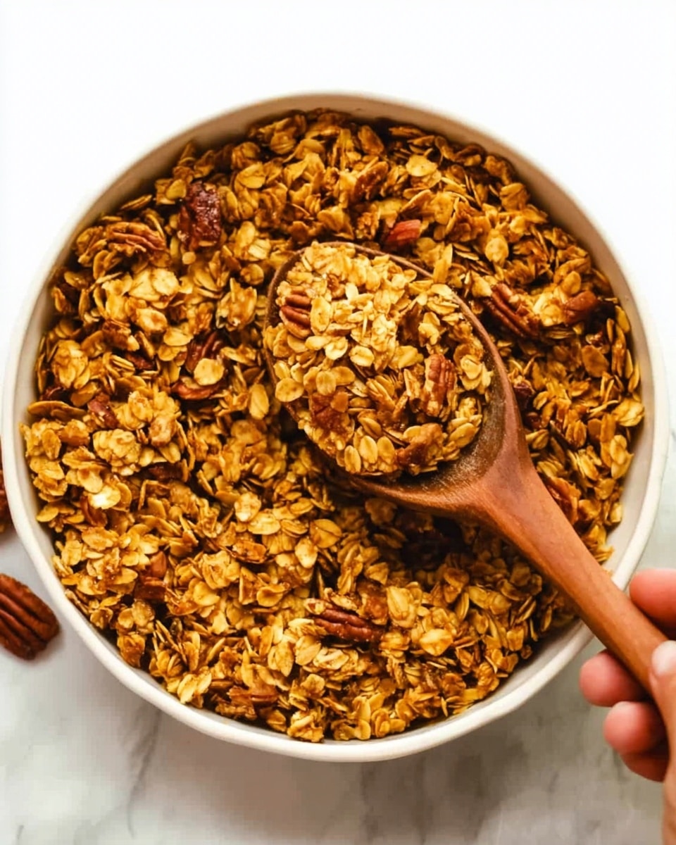 A top-down view of a white bowl filled with golden brown granola. The granola has large oat flakes mixed with small nuts and seeds, creating a rough, crunchy texture. A wooden spoon, held by a woman's hand, is scooping some of the granola from the bowl. The bowl is placed on a white marbled surface. The overall color is warm with tones of brown, tan, and light yellow. photo taken with an iphone --ar 4:5 --v 7