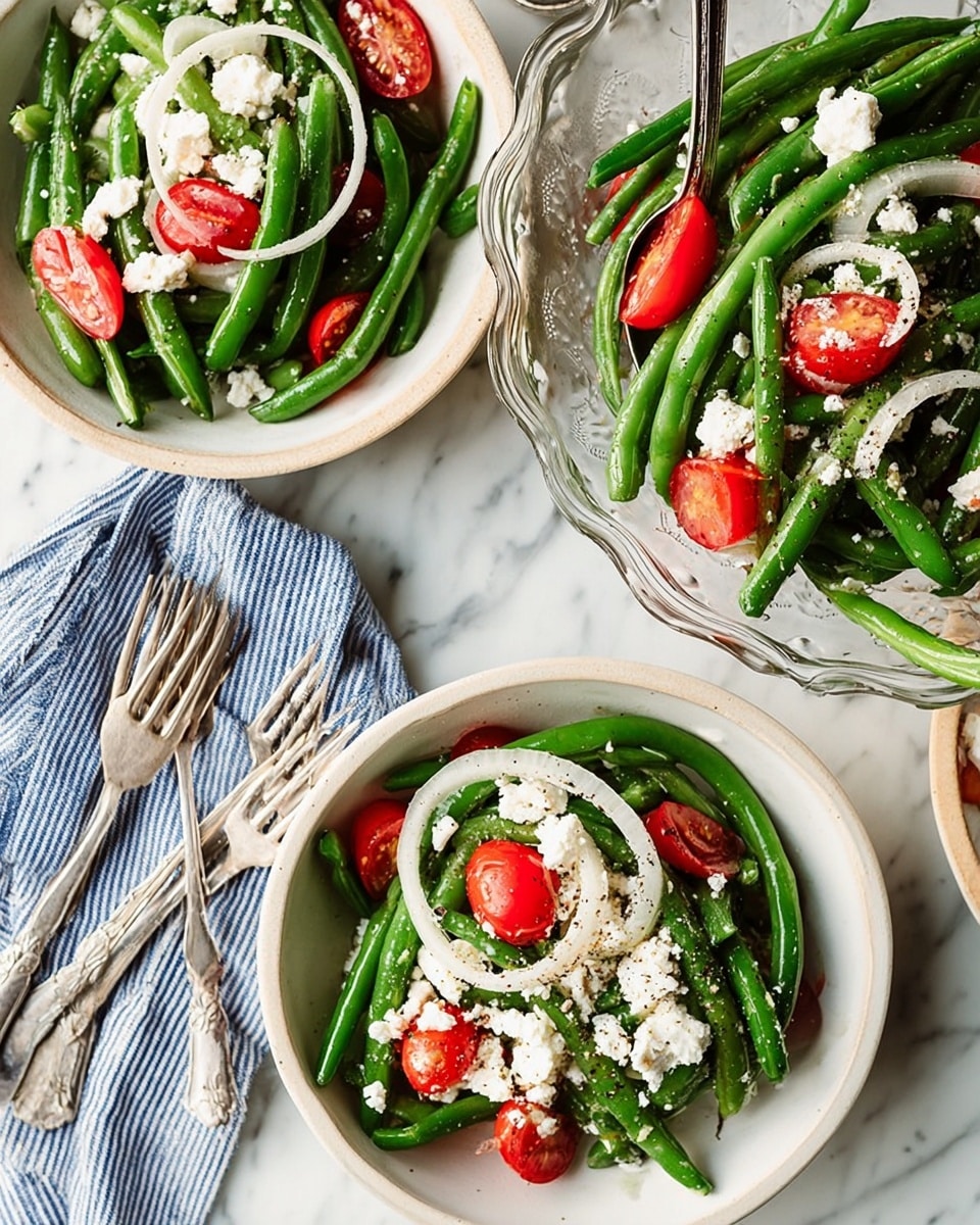 A fresh green bean salad is shown in three white bowls placed on a white marbled surface. Each bowl contains bright green beans as the main layer, with slices of red cherry tomatoes and thin rings of white onions scattered on top. Crumbled white cheese pieces are sprinkled generously over the salad layers, with a light dusting of black pepper visible on the cheese and vegetables. The largest bowl is clear with a decorative edge and has a metal spoon resting inside. Close to the bowls, a striped blue and white cloth napkin lays next to two silver forks. The colors are fresh and vibrant, focused on green, red, and white elements, creating a bright and healthy look photo taken with an iphone --ar 4:5 --v 7