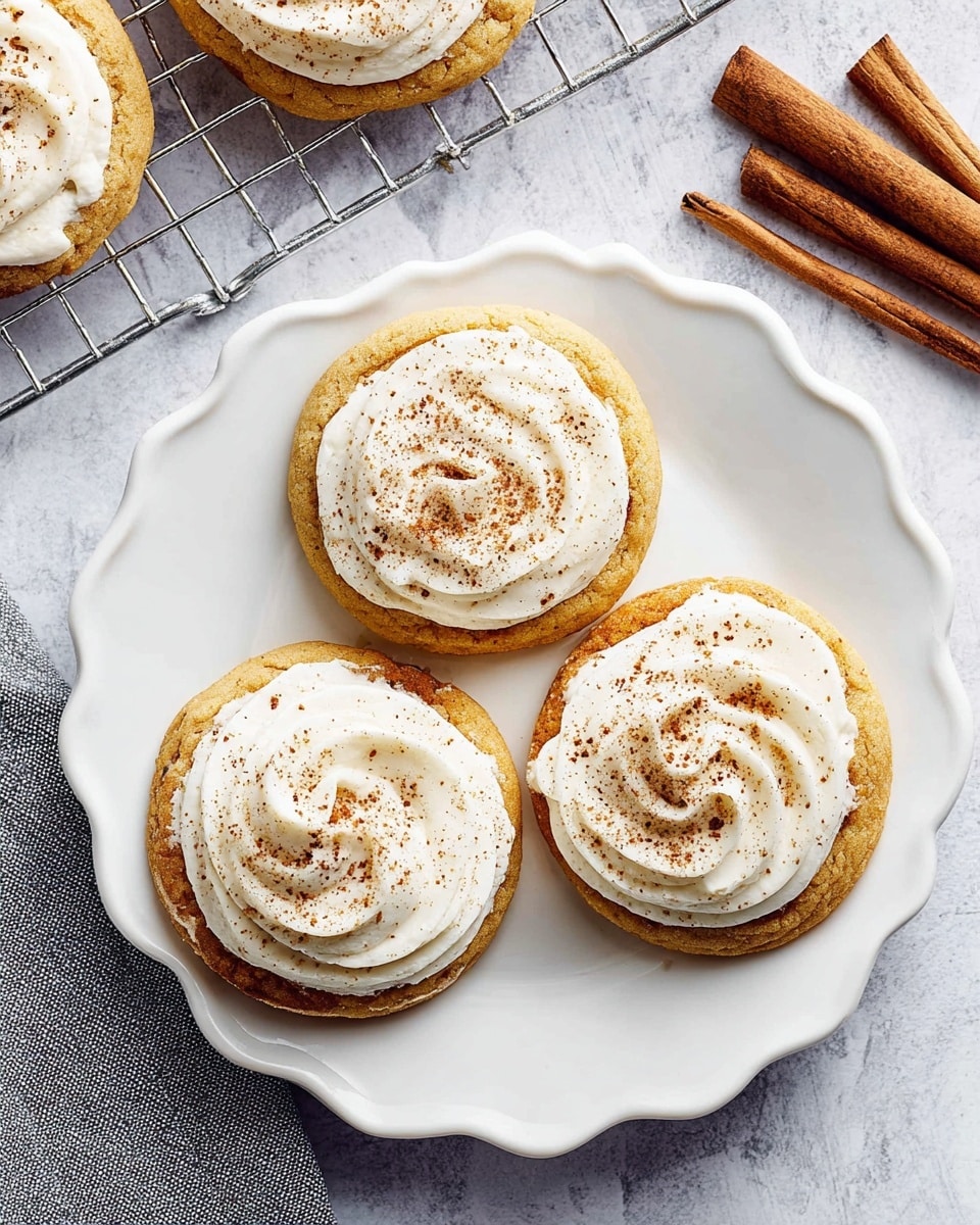 Three round cookies are placed on a white scalloped plate on a white marbled surface. Each cookie has two layers: the bottom layer is a soft golden-brown cookie with a smooth texture, and the top layer is a thick swirl of white frosting sprinkled evenly with light brown cinnamon powder. To the right of the plate are two cinnamon sticks, and above the plate is a metal cooling rack with more cookies, partially visible, and a folded gray cloth. Photo taken with an iphone --ar 4:5 --v 7
