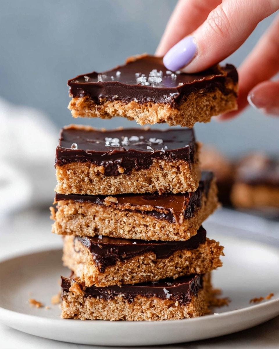 A stack of five square cookie bars with a golden-brown, slightly crumbly texture forms the base, each layer topped with a smooth, dark chocolate layer that has a few shiny salt flakes scattered on top. The layers show a contrast between the light, textured cookie and the glossy chocolate. A woman's hand with light purple nail polish is lifting the top piece, revealing the underside and edges of the chocolate and cookie layers. The bars are sitting on a white plate placed on a white marbled surface, with a blurred background giving focus to the stack. Photo taken with an iphone --ar 4:5 --v 7