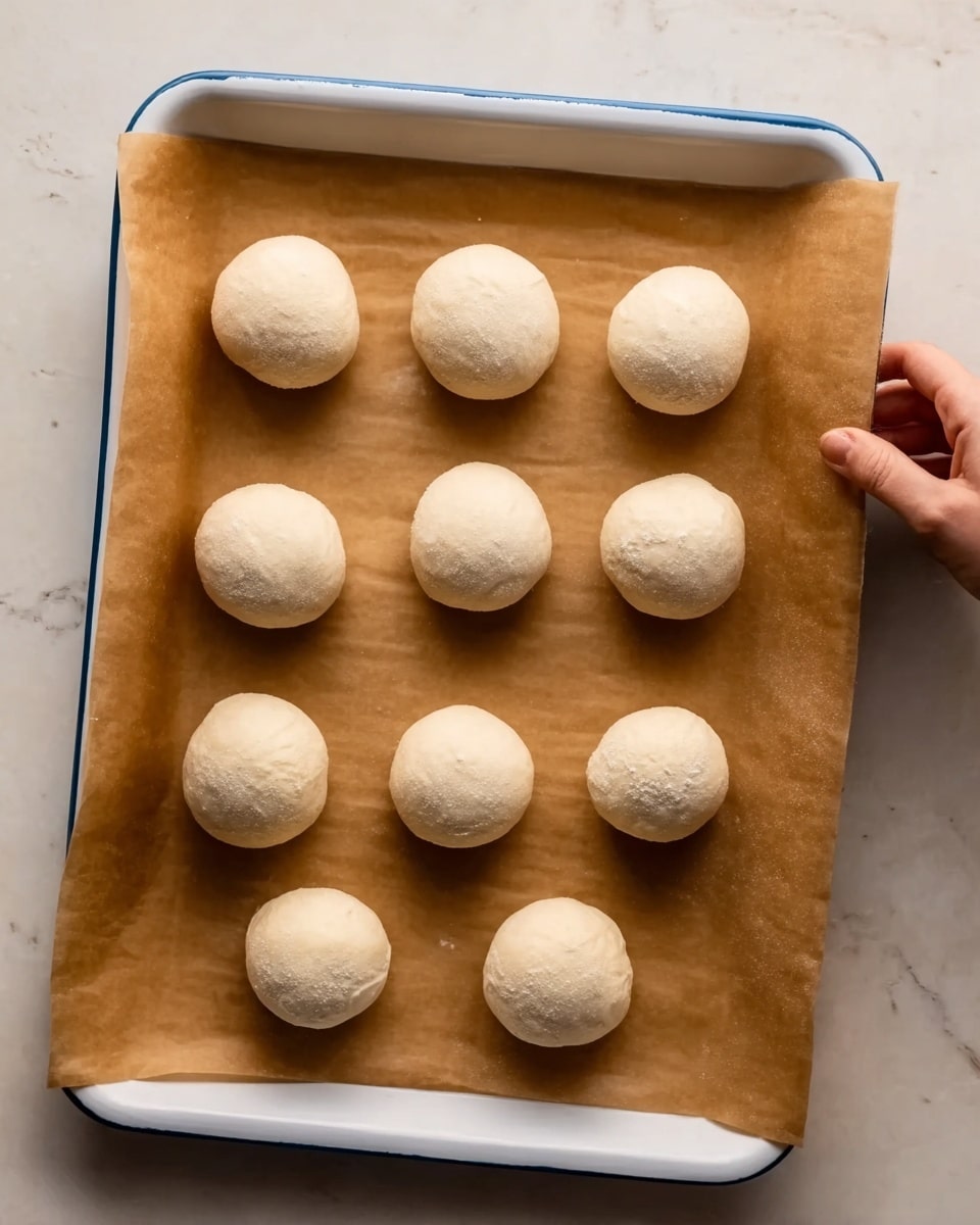 The image shows a white baking tray lined with brown parchment paper, resting on a white marbled surface. On the parchment paper, there are nine evenly spaced dough balls, each smooth and pale beige in color. The dough balls appear soft and round, with slight flour dusting giving them a matte texture. The scene also includes a woman's hand entering from the right side, gently touching or adjusting one of the dough balls. Photo taken with an iphone --ar 4:5 --v 7