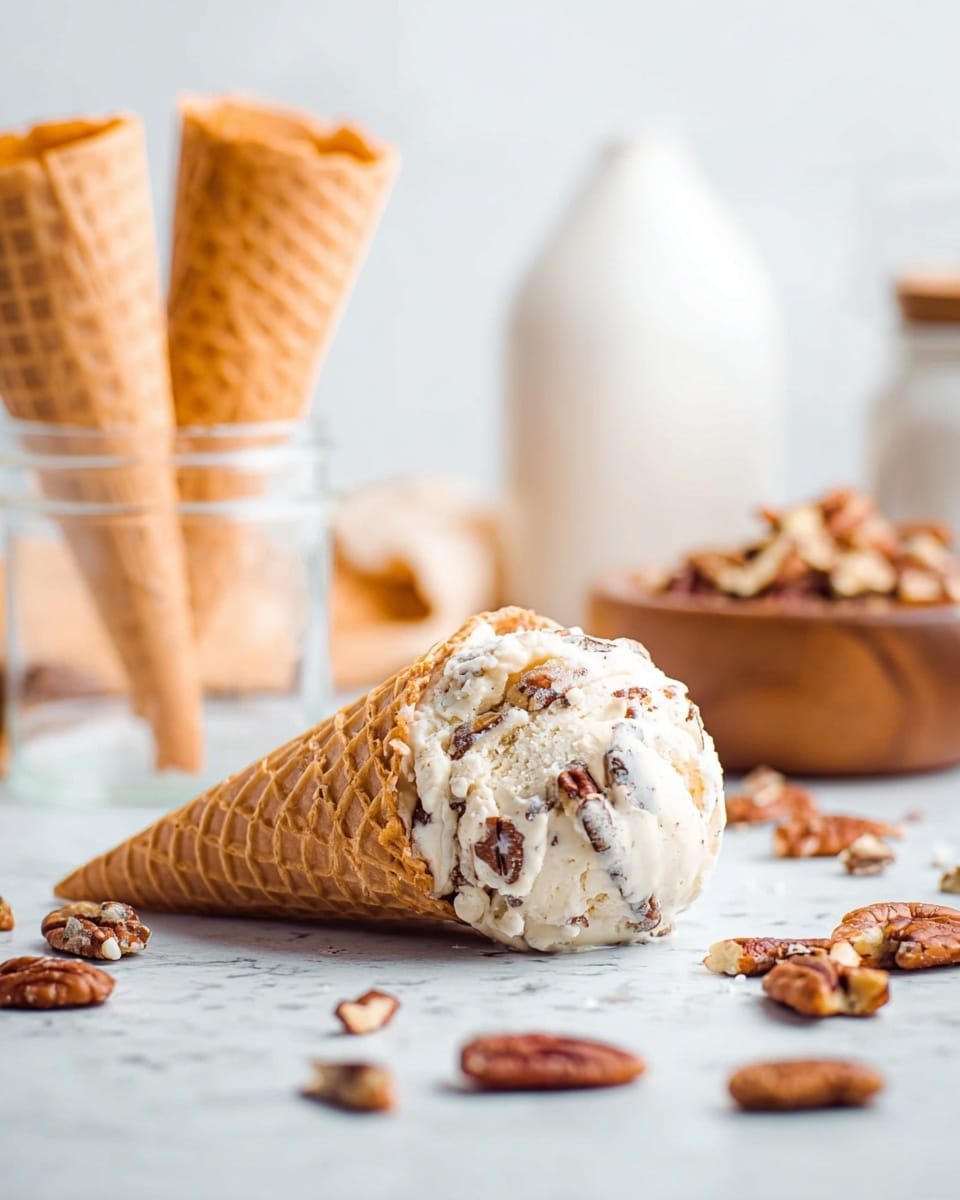 The image shows one waffle cone lying on a white marbled surface, filled with one scoop of ice cream mixed with chunks of brown nuts scattered throughout. Around the cone, there are loose pieces of pecans spread on the white marbled surface. To the left, there are two empty waffle cones placed upright inside a clear glass jar. In the background, there is a small wooden bowl filled with more brown nuts, softly blurred, along with a white ceramic container also out of focus. The whole scene is bright and airy with soft natural light. photo taken with an iphone --ar 4:5 --v 7