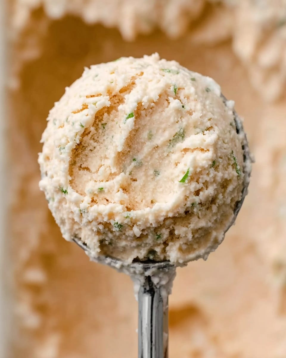 The image shows a close-up of a round scoop of a smooth, creamy mixture with a light beige color and small green flecks inside. The scoop is held by a metal ice cream scooper, and the background is filled with more of the same mixture, matching the texture and color. The scene sits on a white marbled surface. photo taken with an iphone --ar 4:5 --v 7