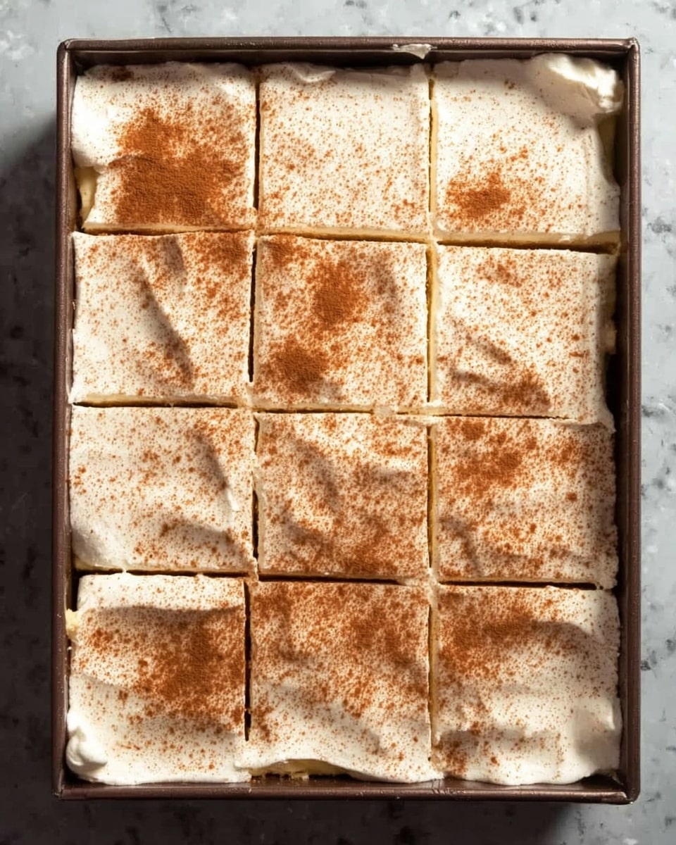 The image shows a rectangular dark baking pan filled with a dessert cut into 12 square pieces. The top layer is a thick, fluffy white cream sprinkled evenly with brown cinnamon powder. The cream layer looks soft and smooth with slight cracks on some pieces. The dessert rests on a white marbled surface. photo taken with an iphone --ar 4:5 --v 7