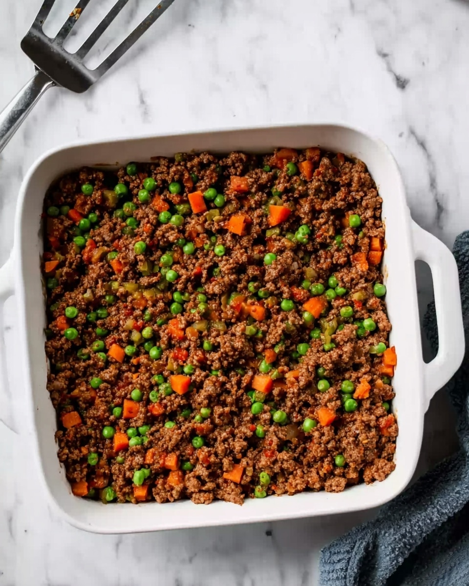 A square white baking dish filled with a thick layer of cooked ground meat mixed with bright green peas and small orange carrot cubes evenly spread across the top. The mixture has a slightly rough, chunky texture with a rich brown color from the meat, contrasted by the vibrant vegetables scattered throughout. The dish is placed on a white marbled surface, and a metal spatula with a black handle rests nearby. photo taken with an iphone --ar 4:5 --v 7