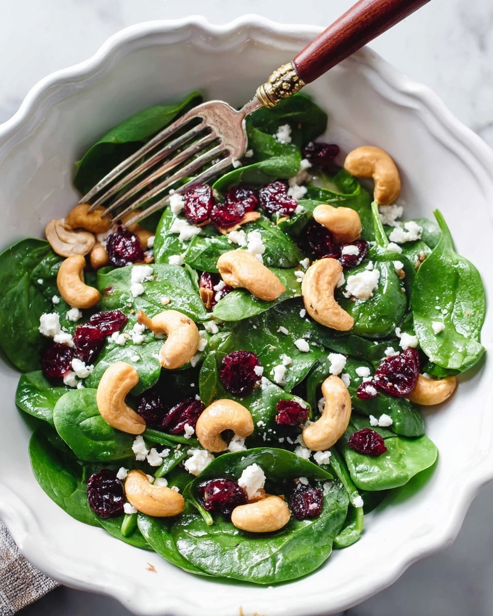 A white scalloped plate filled with fresh green spinach leaves as the base layer, topped with whole light brown cashew nuts scattered evenly, bright red dried cranberries placed over the cashews and spinach, and small white crumbles of cheese sprinkled across the top. Tiny black chia seeds are spread lightly over all layers. The plate sits on a white marbled surface. photo taken with an iphone --ar 4:5 --v 7
