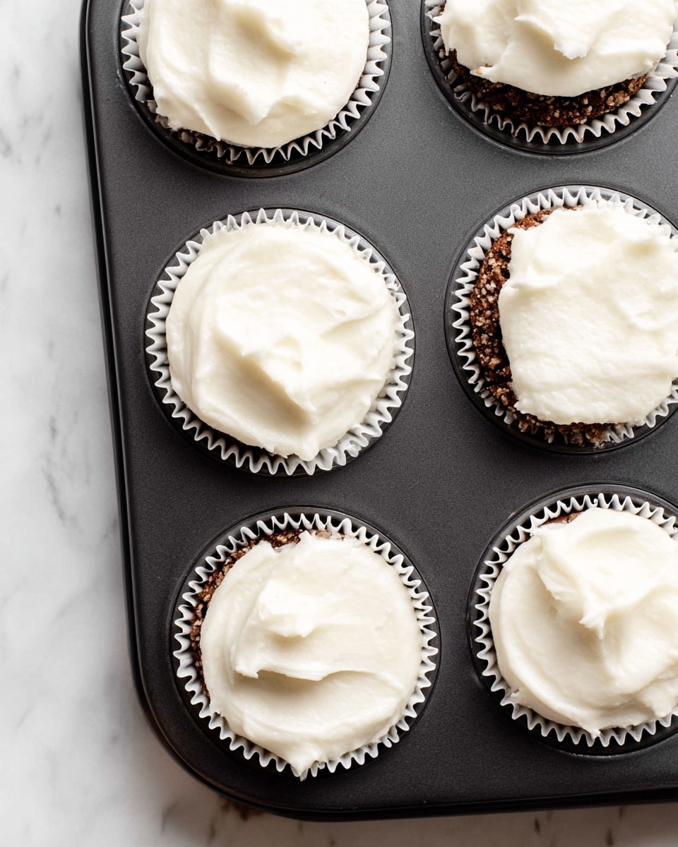 A close-up view of a black muffin tray holding six cupcakes, each in a white paper liner. The cupcakes have two visible layers: the bottom layer is a dark, crumbly base, and the top layer is a thick, smooth, white frosting that covers the surface of each cupcake unevenly with soft peaks and swirls. The tray rests on a white marbled surface, and the image is bright with soft, natural lighting, creating a clean, fresh look. photo taken with an iphone --ar 4:5 --v 7