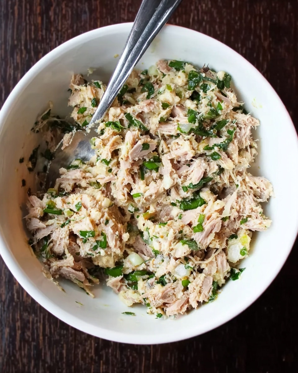 The image shows a white bowl filled with a mix of shredded beige and light brown tuna pieces combined with small green leafy bits and tiny white pieces, giving a slightly chunky texture. A silver spoon is inside the bowl resting on the left edge, partially covered with the tuna mix. The bowl sits on a dark wooden surface. The overall look is creamy but not smooth, with visible bits of green herbs spread throughout. photo taken with an iphone --ar 4:5 --v 7