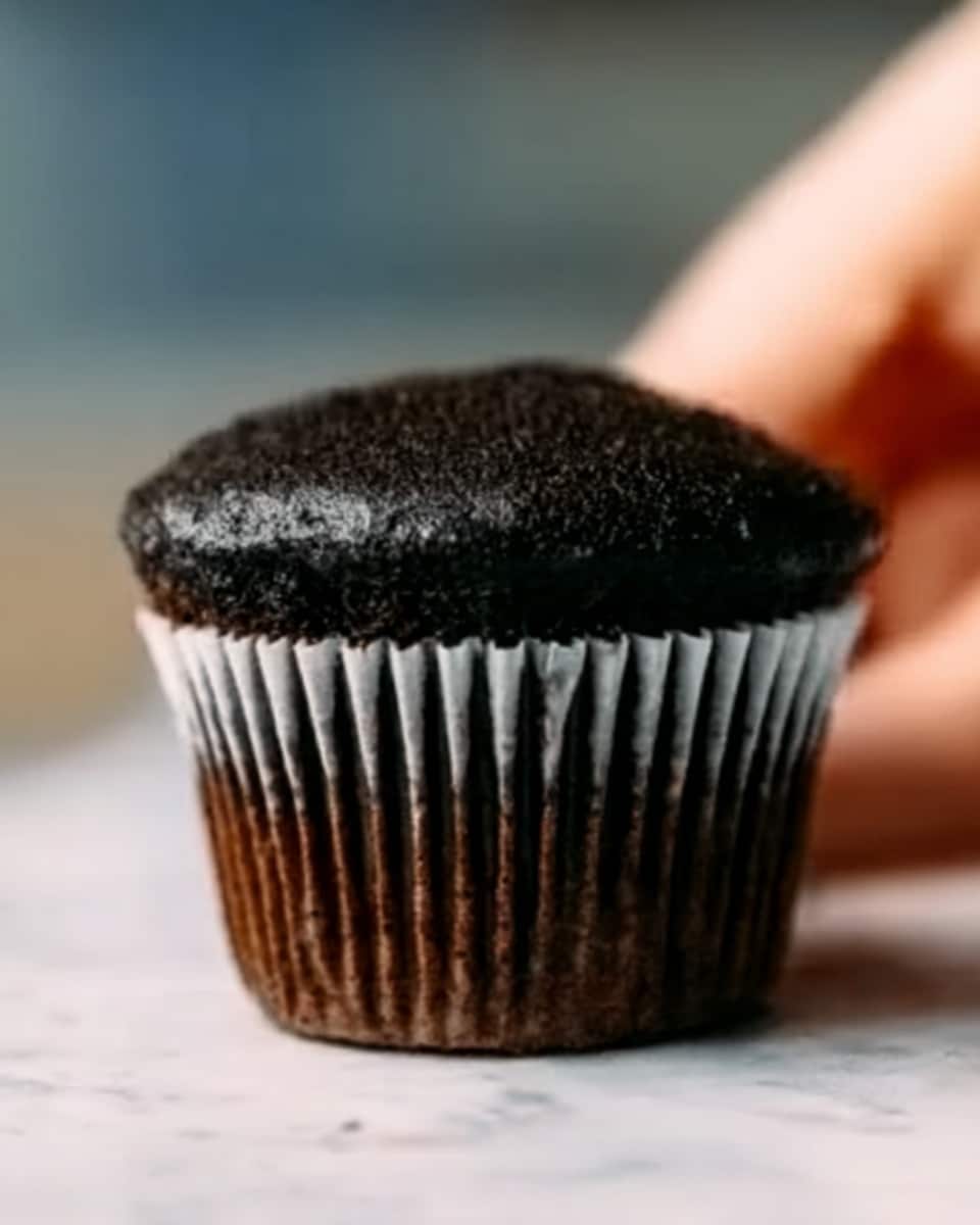 A close-up of a chocolate cupcake with a dark brown base and a thick top layer of smooth, black frosting, both inside a white cupcake liner. The cupcake is placed on a white marbled surface, with a blurred background showing part of a woman's hand holding it. The overall look is simple and rich in dark tones, focused on the texture and color of the cupcake. photo taken with an iphone --ar 4:5 --v 7