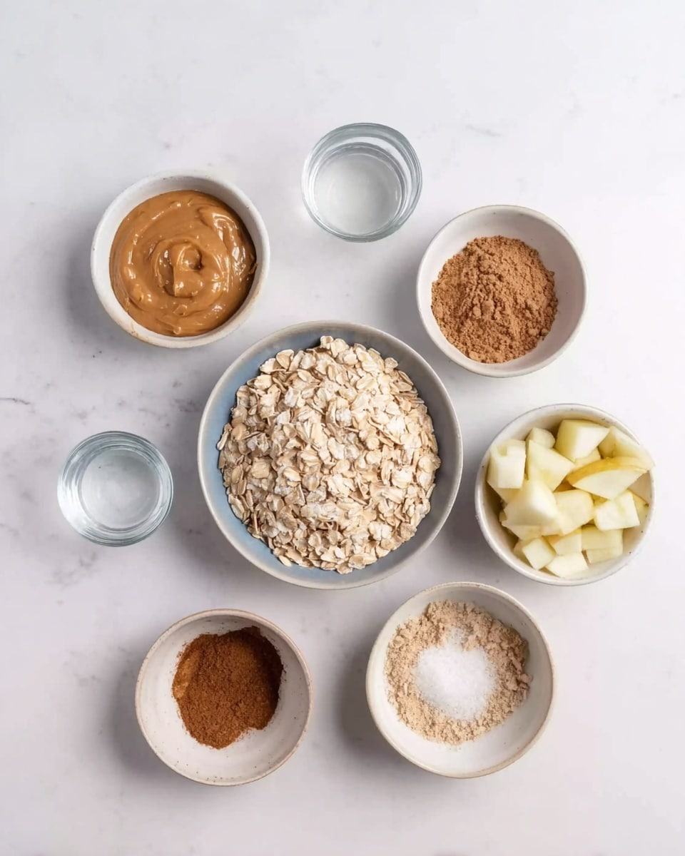 On a white marbled surface, there are seven small white bowls arranged loosely in a circle. In the center is a bowl filled with light beige rolled oats with a bumpy texture. Surrounding it, starting from top and moving clockwise, are a bowl of smooth, light brown peanut butter, a bowl with fine brown powder, a transparent plastic cup of clear water, a bowl with coarse medium brown powder, a tiny bowl with fine white salt, and a bowl with small, pale yellow apple cubes. The colors mostly range from light beige to various shades of brown and pale yellow. Photo taken with an iphone --ar 4:5 --v 7