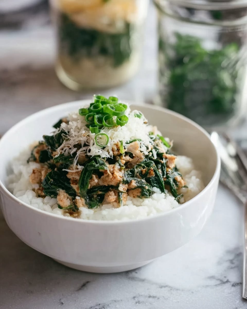A white bowl sits on a white marbled surface filled with a base layer of soft, white rice. On top of the rice is a layer of cooked dark green spinach mixed with light brown pieces of tofu or a similar plant-based protein. Over this, there is a small pile of thinly shredded white cheese or radish, and the dish is finished with a sprinkle of bright green chopped scallions placed in the center. In the background, there are blurred images of glass jars with layers of similar ingredients. The lighting is soft and natural, highlighting the fresh colors of the dish. Photo taken with an iphone --ar 4:5 --v 7