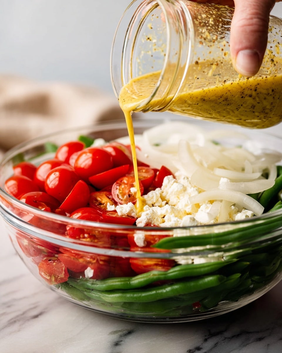 A clear glass bowl filled with three visible layers: the bottom layer is made of green beans, the middle layer holds a mix of halved bright red cherry tomatoes, and the top layer includes crumbled white cheese and sliced white onions. A woman's hand is pouring a thick yellow dressing, flecked with black pepper, from a clear jar over the ingredients. The bowl is placed on a white marbled surface with a soft blurry background. photo taken with an iphone --ar 4:5 --v 7