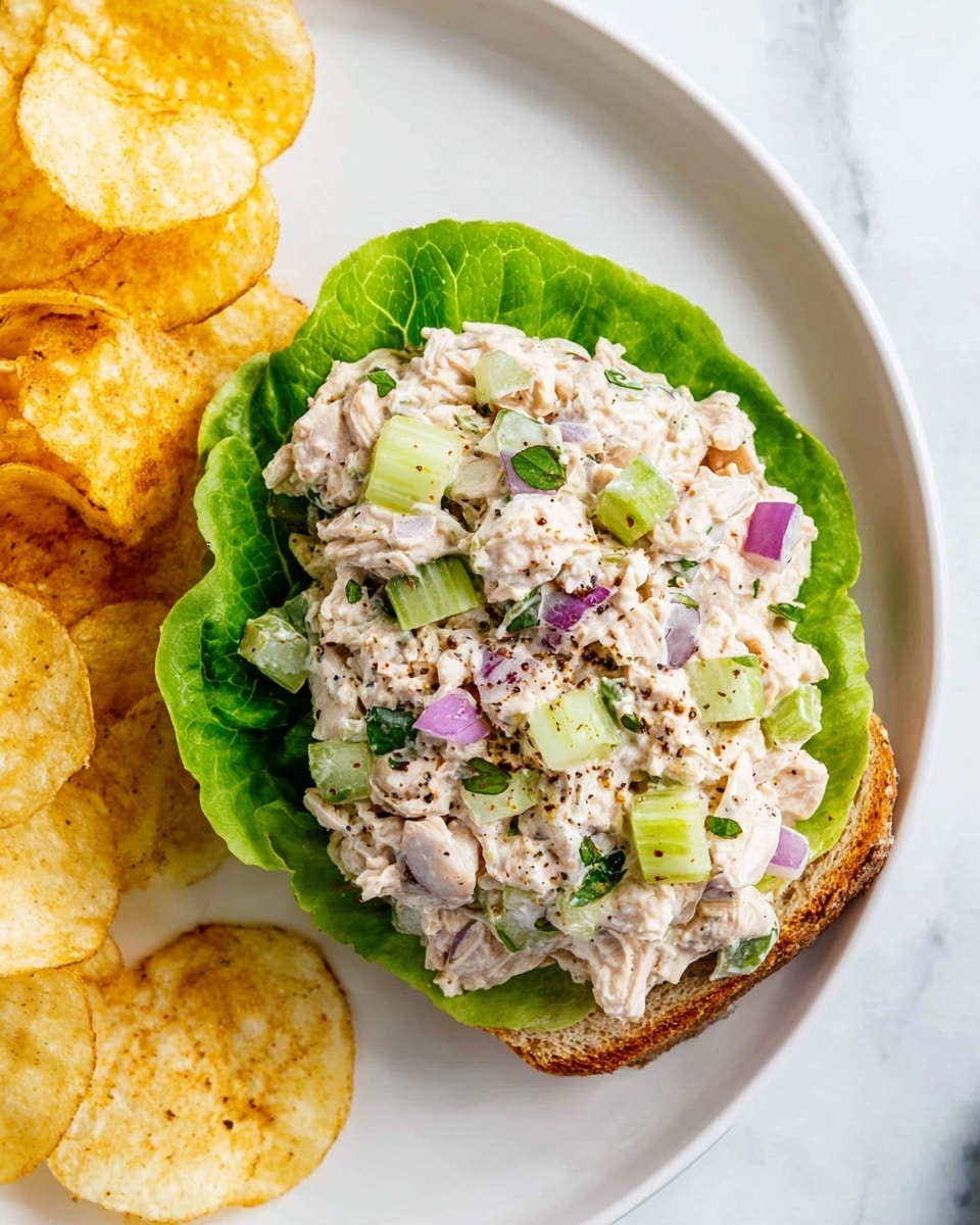 A single slice of toasted whole grain bread sits on a white plate with some black pepper and spice specks scattered around. The bottom layer on the bread is a fresh, bright green lettuce leaf, which has a smooth and slightly wavy texture. On top of the lettuce, there is a thick, uneven layer of creamy tuna salad mixed with small chunks of light beige tuna, pieces of green celery, and tiny bits of purple-red onion, all speckled with black pepper. The colors range from light cream to green and purple, creating a fresh and textured look. The scene is set on a white marbled surface. photo taken with an iphone --ar 4:5 --v 7