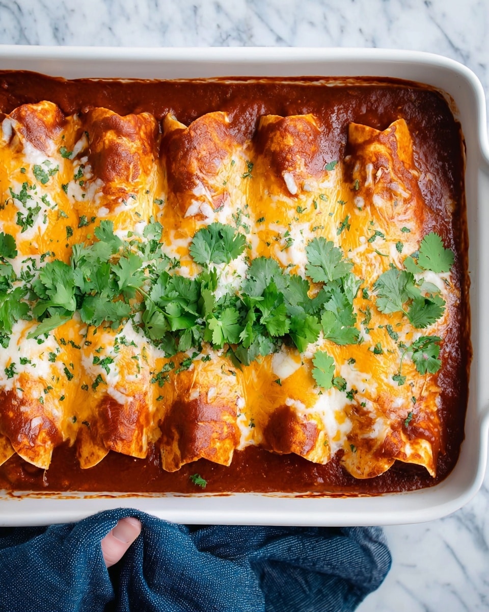 The dish is a white baking dish filled with five rolled tortillas placed side by side, each covered with melted orange and white cheese and a thick layer of red sauce that slightly bubbles and browns on top. The tortillas show some soft texture with cheese melting around them, sprinkled lightly with chopped green herbs, and topped with fresh green cilantro leaves spread across the top. The dish sits on a white marbled surface and is being held by a woman's hand with a dark blue cloth. Photo taken with an iphone --ar 4:5 --v 7