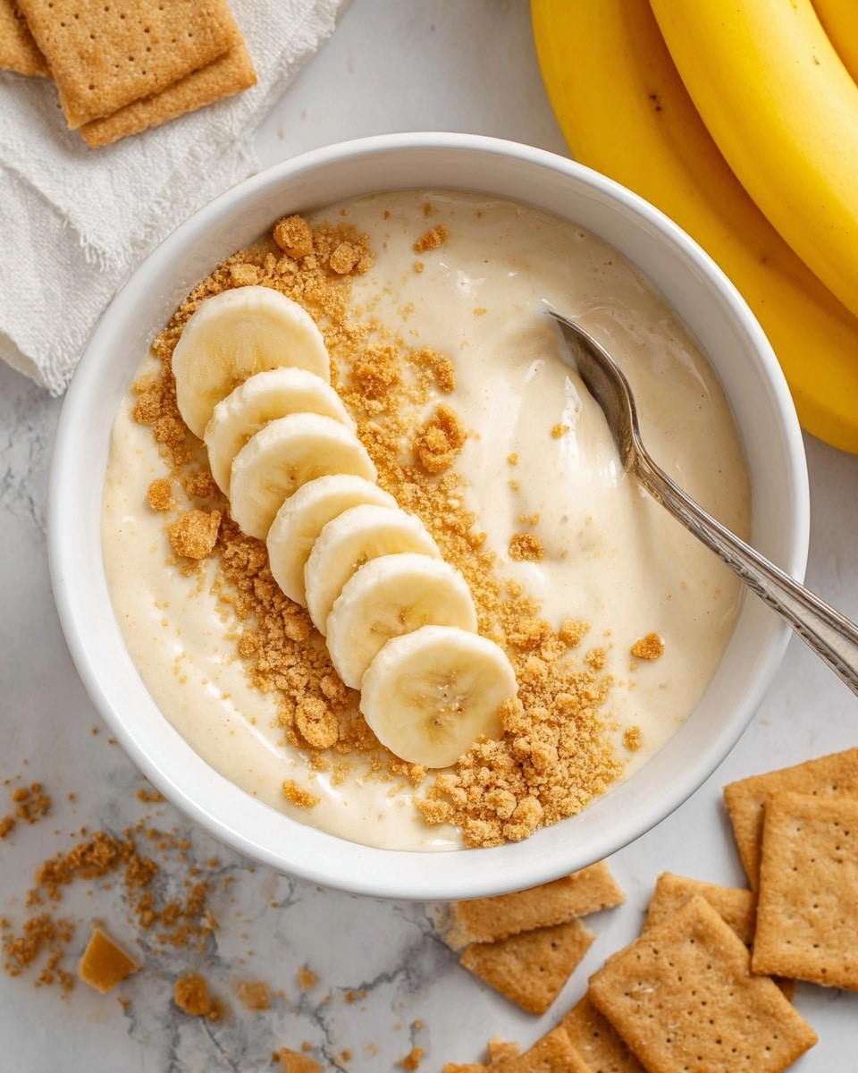 A white bowl filled with creamy beige pudding as the base layer, topped with a neat line of five round banana slices slightly overlapping on the right side. Crushed light brown cookie crumbs are sprinkled over the pudding evenly, adding texture. A shiny silver spoon is partially dipped into the pudding on the left side. The bowl sits on a white marbled surface, with whole and broken pieces of light brown crackers scattered near the bottom right. A bunch of yellow bananas is seen on the top right edge of the image. Photo taken with an iphone --ar 4:5 --v 7