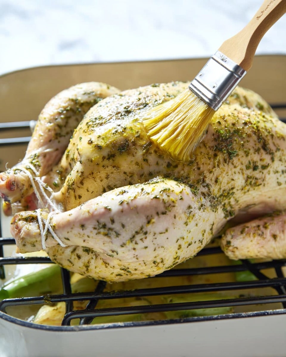 A whole raw chicken lies on a black metal rack inside a white roasting pan, with green herbs and spices rubbed all over its skin giving it a speckled light green and yellow look. A brush with a wooden handle is seen applying a layer of yellow oil or butter sauce on the chicken's top, adding a shiny texture. The chicken is tied at the legs with white kitchen string, and underneath the rack some light green vegetables are slightly visible. The background surface is white marble and the scene is bright, showing natural light on the chicken photo taken with an iphone --ar 4:5 --v 7