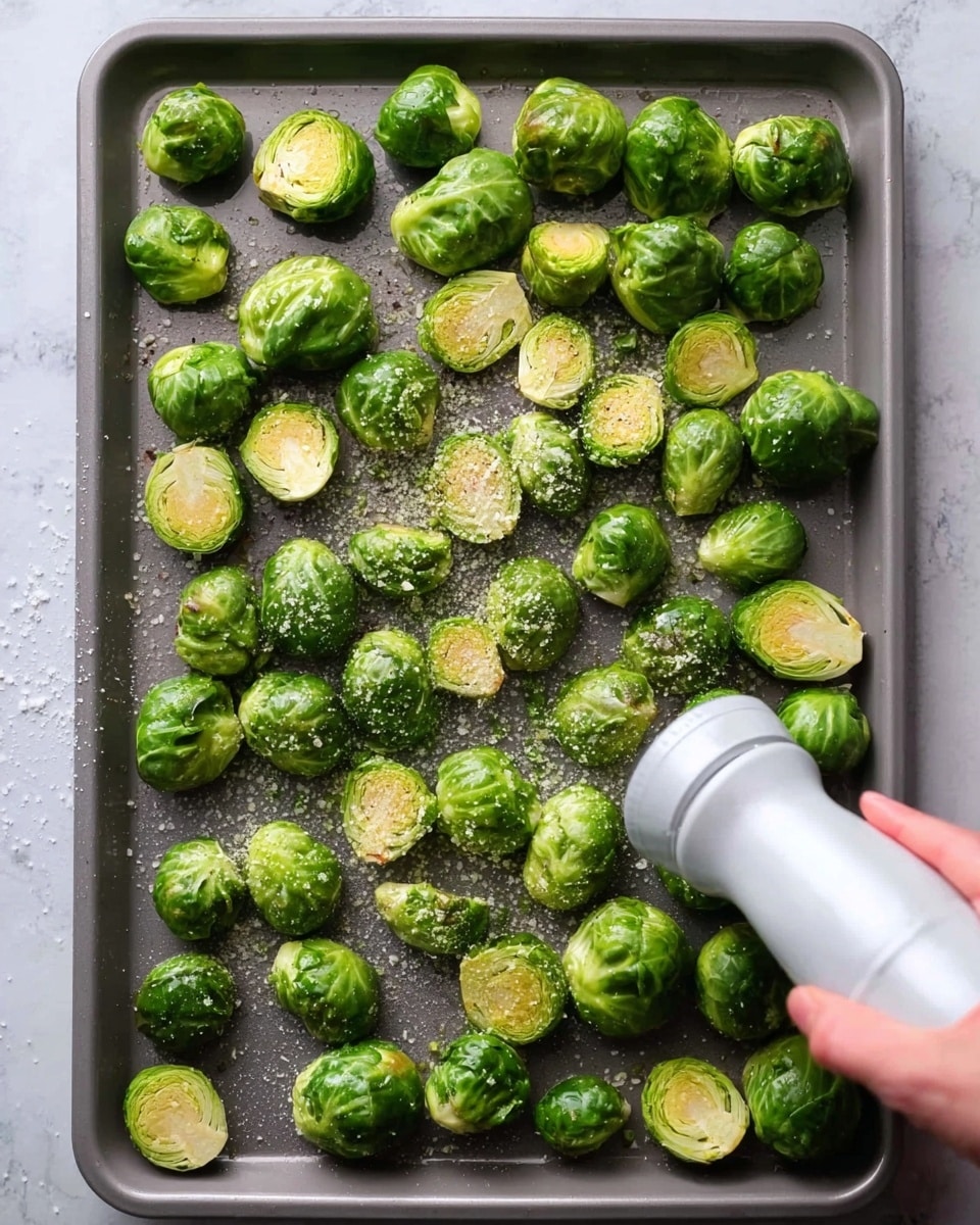 The image shows a gray baking tray filled with whole and halved bright green Brussels sprouts spread out evenly. The Brussels sprouts are coated lightly with oil, and there is coarse salt and black pepper sprinkled across them. In the bottom right corner, a woman's hand holds a white pepper grinder above the tray. The background is a white marbled texture. photo taken with an iphone --ar 4:5 --v 7
