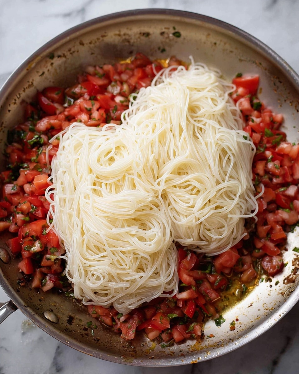 A metal pan filled with diced red tomatoes and green herbs scattered at the bottom, topped with two large piles of white, thin noodles that have a smooth and slightly shiny texture, sitting neatly in the center of the pan. The tomatoes and herbs create a colorful, chunky base layer under the pale, smooth noodles. The pan surface shows small spots of cooking juices around the edges, and the overall background is a white marbled texture. photo taken with an iphone --ar 4:5 --v 7
