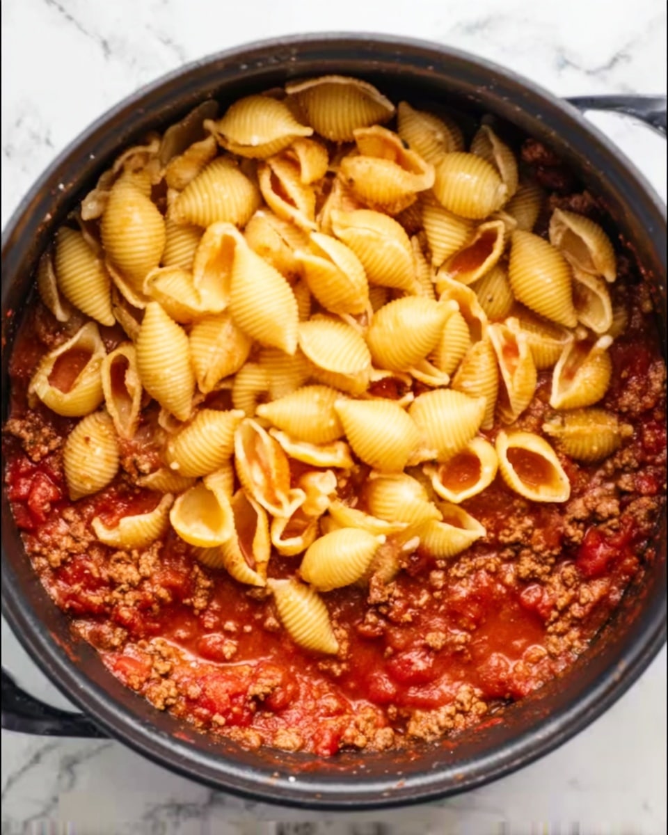 A black pot filled with partially cooked yellow pasta shells piled on top, resting above a rich red tomato sauce with visible ground meat mixed in. The sauce has a slightly chunky texture with bits of tomato scattered around the edges. The pot is sitting on a white marbled surface, and the image is taken from a top-down view showing all the layers clearly. Photo taken with an iphone --ar 4:5 --v 7