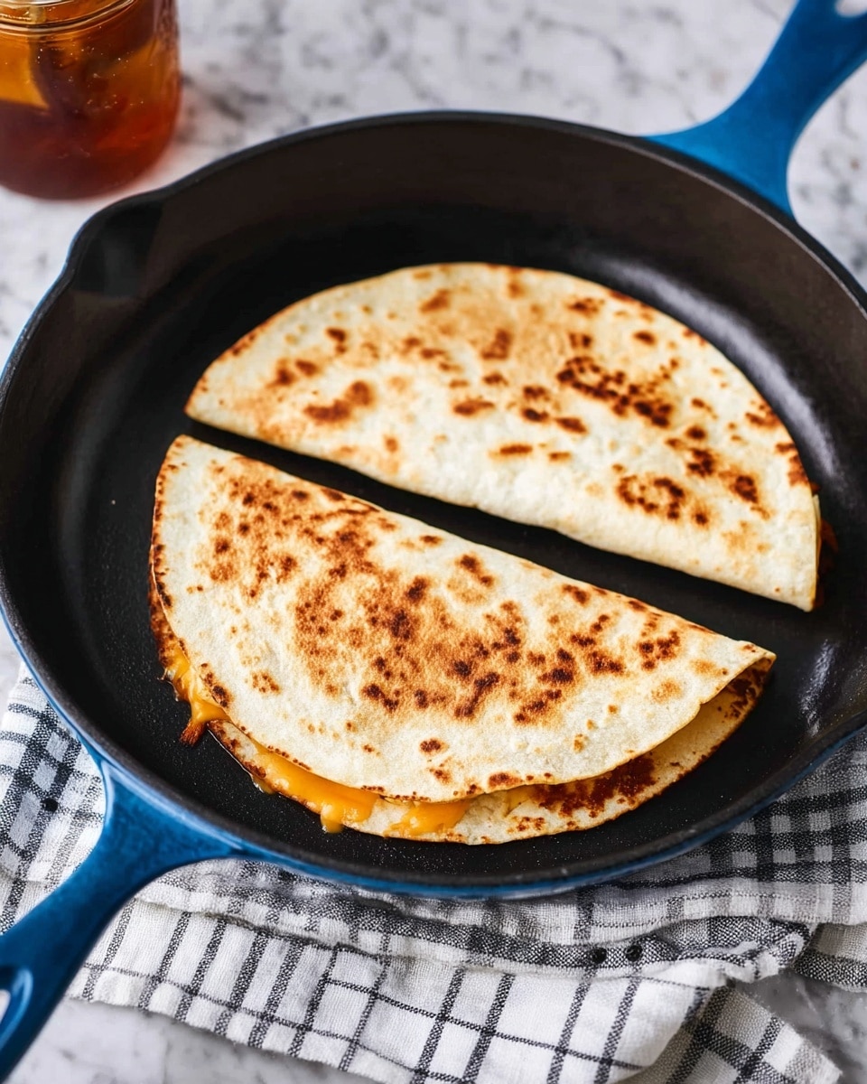 Two half-moon shaped quesadillas with a golden brown toasted texture on top lie side by side inside a black cast iron pan with a blue handle. The quesadillas are folded tortillas with a slightly crispy surface showing darker brown spots from cooking. A small amount of melted orange cheese peeks out from the edge of one quesadilla. The pan is placed on a white and black checkered cloth that rests on a white marbled surface. A glass jar with iced tea is blurred in the background. Photo taken with an iphone --ar 4:5 --v 7