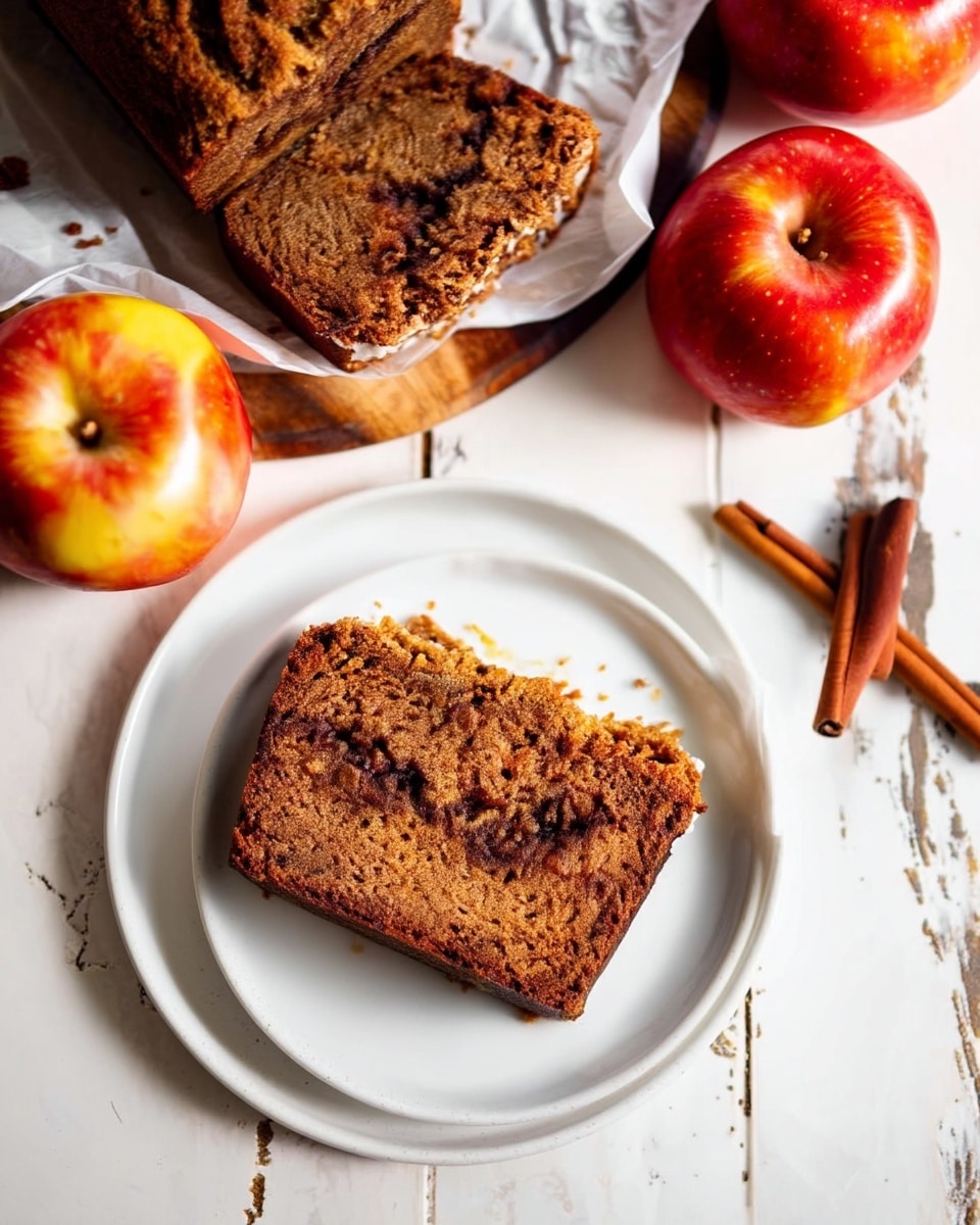 The image shows a single slice of dark brown spiced cake with a moist texture and a visible darker swirl layer near the middle, sitting centered on a white round plate. Above the plate is a wooden board lined with white crinkled parchment paper holding the remaining loaf of the same cake, which has a rough and soft crumb texture. Around the plate and board are three red and yellow apples with shiny skins, and to the right side are two cinnamon sticks. The background surface is a white marbled texture with slight rustic marks. Photo taken with an iphone --ar 4:5 --v 7