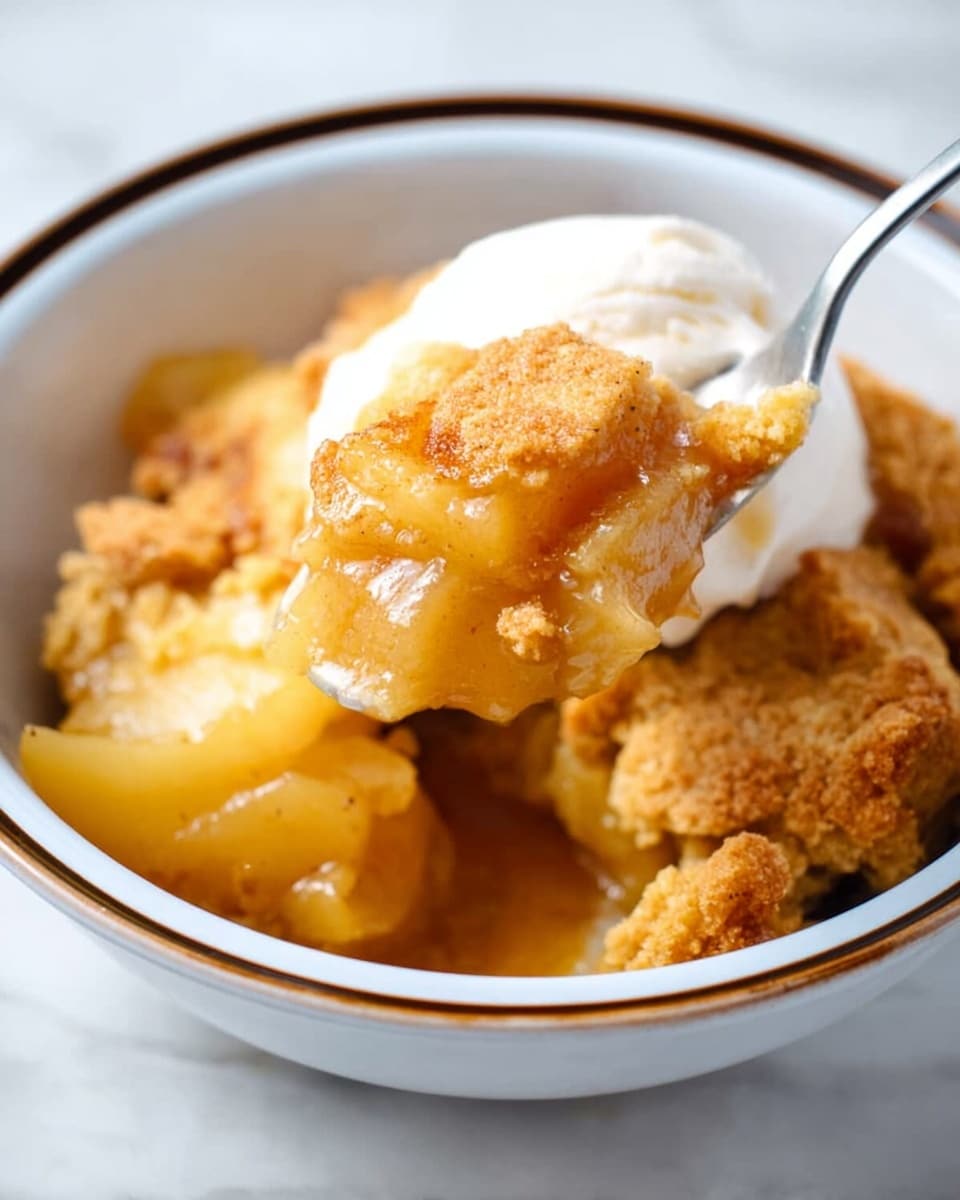 A close-up of a spoon lifting a portion of apple cobbler from a white bowl with a dark rim, showing two main layers: the bottom layer is soft, golden apple slices with syrup soaking the area, and the top layer is a crumbly, golden-brown biscuit crust that looks crunchy and thick. On the side inside the bowl, there is a smooth, white scoop of ice cream melting slightly into the cobbler, creating a mix of creamy and warm textures. The bowl sits on a white marbled surface, with soft lighting enhancing the warm tones of the dessert. Photo taken with an iphone --ar 4:5 --v 7