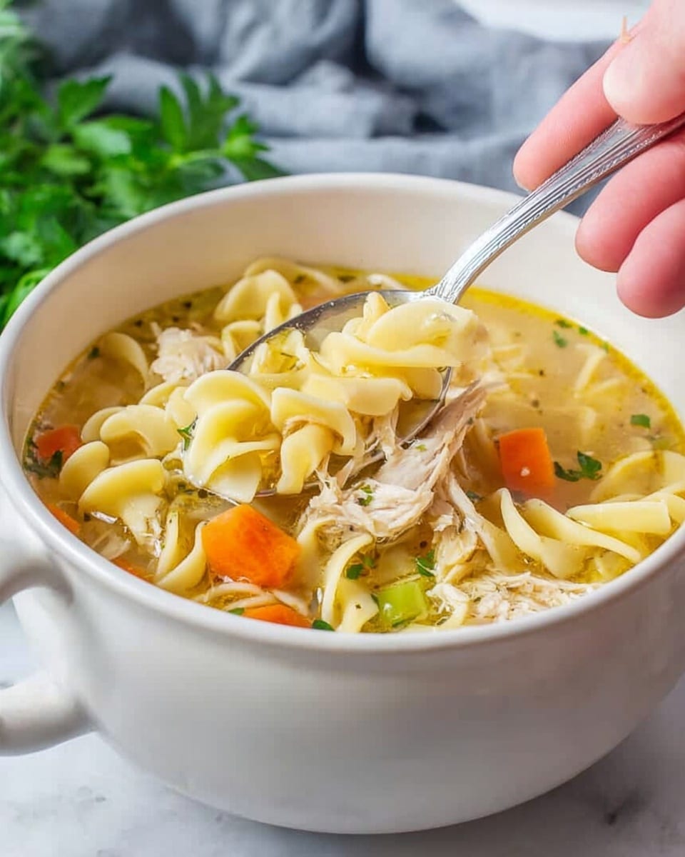 A white ceramic bowl filled with chicken noodle soup sits on a white marbled surface. The soup has three layers visible: the top layer with twisted yellow egg noodles, the middle layer with shredded pale chicken pieces mixed with small bright orange carrot slices and green celery bits, and the bottom layer of clear golden broth. A woman's hand is holding a silver spoon scooping up noodles and chicken from the bowl. Fresh green parsley is seen in the background. The light is soft and natural, highlighting the textures of the noodles and vegetables. photo taken with an iphone --ar 4:5 --v 7