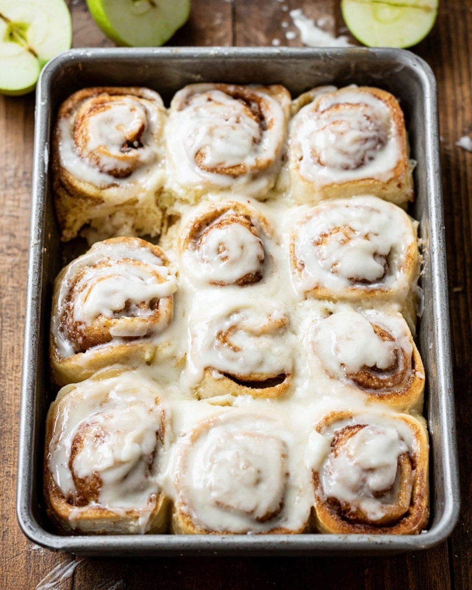 The image shows a metal rectangular baking pan filled with nine cinnamon rolls arranged in three rows of three. Each cinnamon roll has multiple light brown dough layers swirled with darker brown cinnamon filling and is topped with a thick layer of white creamy icing that covers most of the rolls, dripping slightly down the sides. The baking pan is on a wooden surface with two green apple slices in the background. The photo taken with an iphone --ar 4:5 --v 7