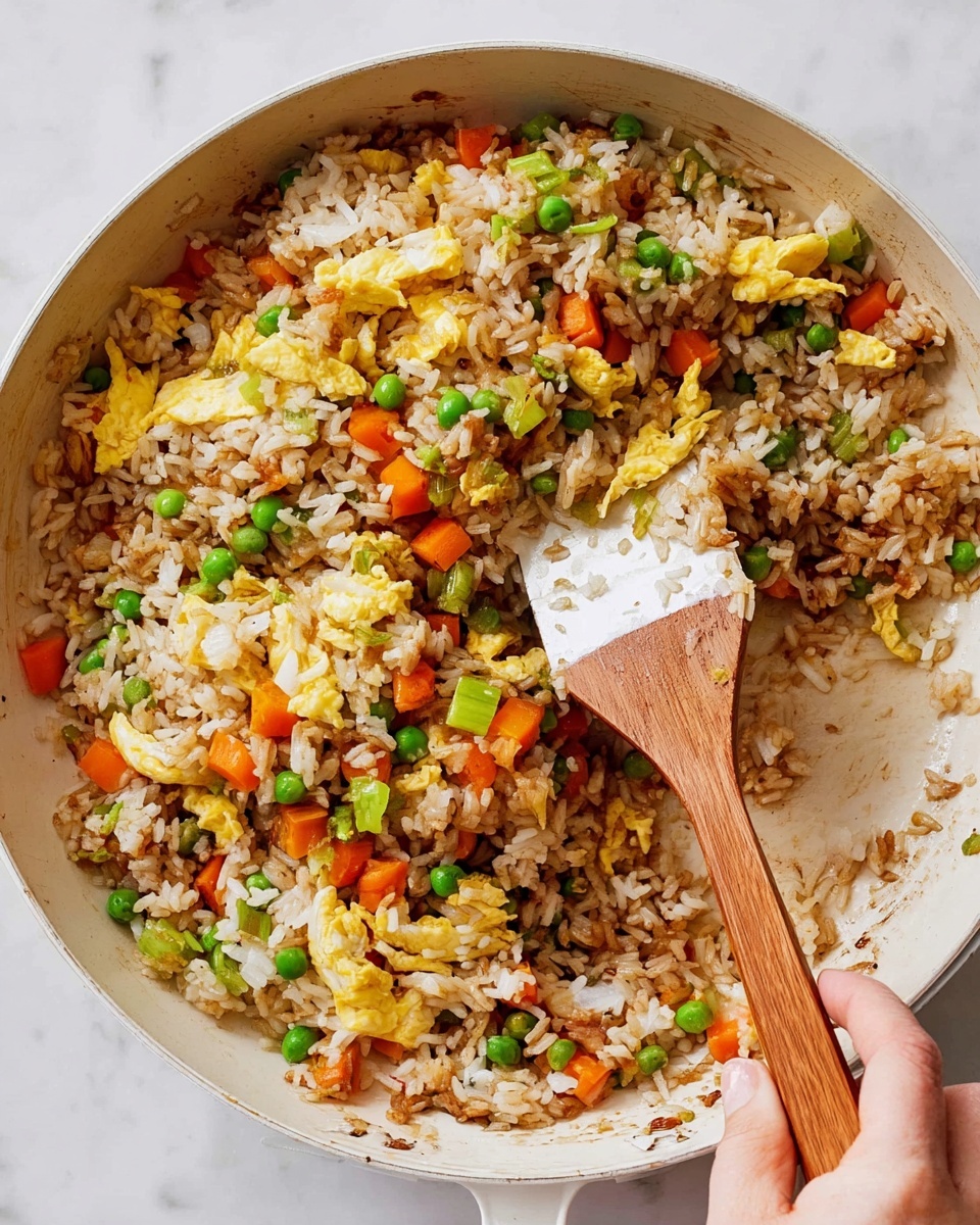 A white pan filled with fried rice showing a mix of white rice grains and brown soy sauce-stained rice grains at the base, scattered with bright orange carrot cubes, green peas, and light green sliced celery. On top, yellow scrambled egg pieces add texture and color contrast. A woman's hand is holding a wooden spatula with a white mixing end, lifting some rice and vegetables from the middle right side of the pan. The background features a white marbled surface, giving a clean look to the scene. photo taken with an iphone --ar 4:5 --v 7