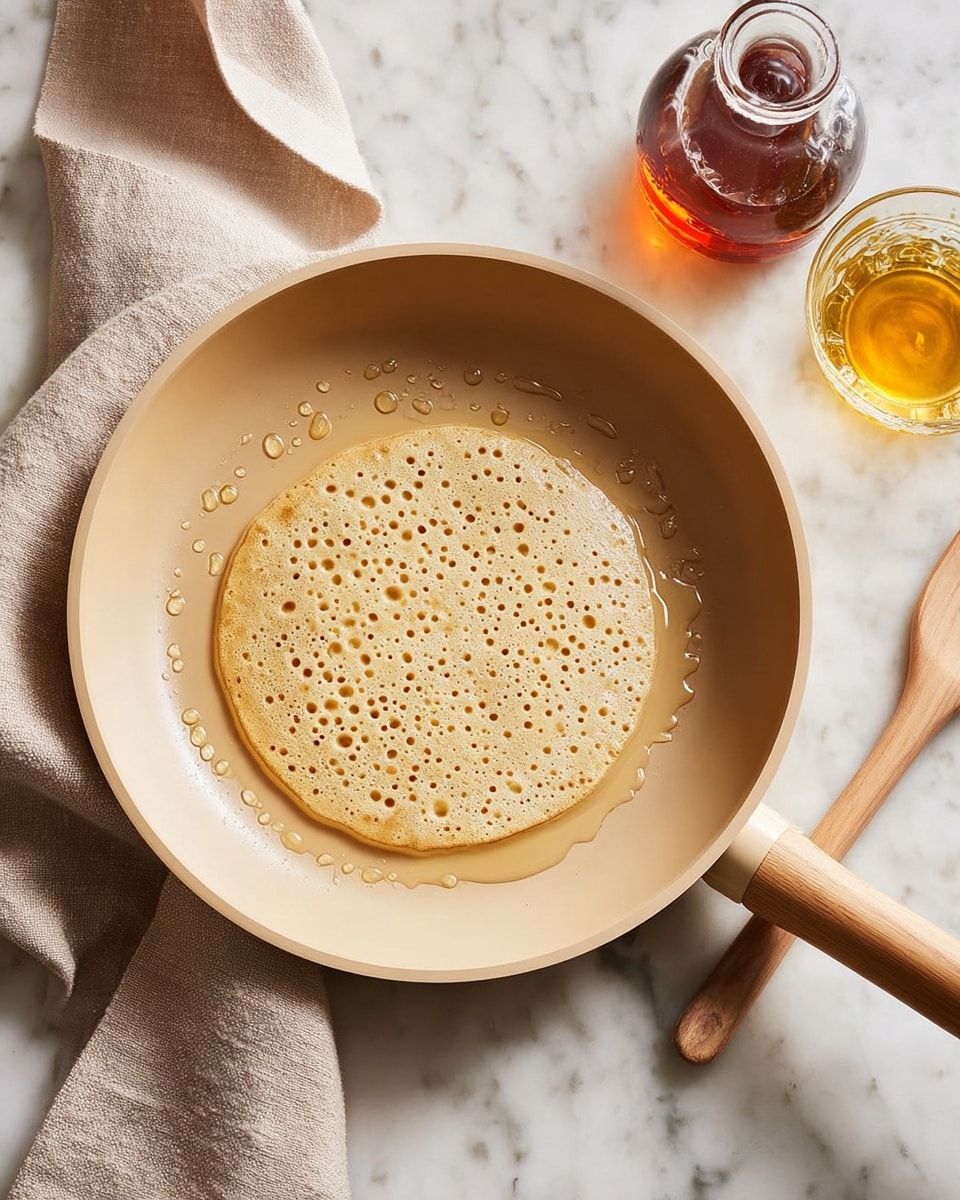 A light tan pancake with many small holes on its surface is cooking in a beige non-stick pan with a wooden handle. The pancake sits in the center of the pan, which has some drops of oil around it. Next to the pan, there is a glass of golden syrup and a glass bottle with dark amber syrup on a white marbled surface. A beige spatula rests at the top right inside the pan, and a beige cloth is placed under the pan handle on the left side. Photo taken with an iphone --ar 4:5 --v 7