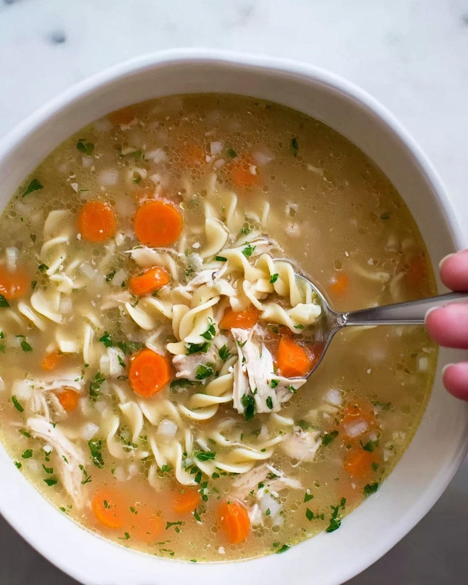 The image shows a close-up of a white bowl filled with chicken noodle soup. The soup has a light, clear broth with visible small diced onions floating on the surface. There are wide egg noodles twisted softly throughout the bowl, mixed with pieces of shredded white chicken and sliced orange carrots. Fresh green herbs are sprinkled lightly over the soup, adding color contrast. A silver spoon is inside the bowl, held by a woman's hand, with some noodles, chicken, and carrot pieces on it. The bowl sits on a white marbled surface. Photo taken with an iphone --ar 4:5 --v 7