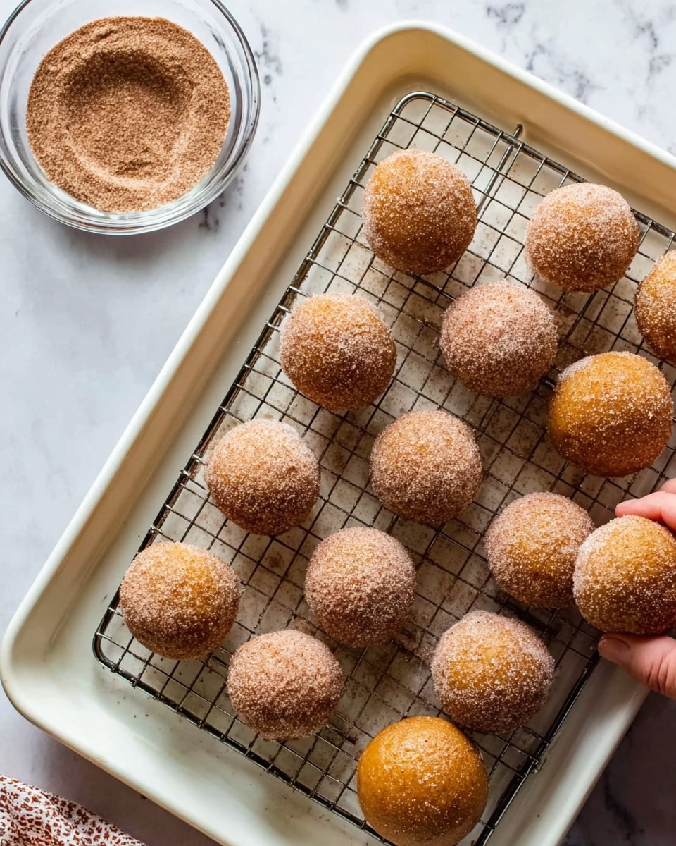 The image shows a white baking tray lined with a cooling rack holding round, golden-brown dough balls covered evenly in a fine layer of cinnamon sugar. In the top part of the tray, there is a small clear glass bowl filled with more cinnamon sugar. A woman's hand is reaching from the top right corner, gently holding one dough ball. The background is a white marbled surface. photo taken with an iphone --ar 4:5 --v 7