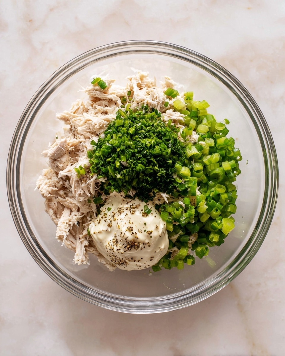 A clear glass bowl shows four main layers: a light beige shredded chicken base spread all around at the bottom, bright green chopped celery and scallions on the right side, a bright green pile of chopped herbs in the center, and a creamy off-white dollop of mayonnaise with specks of black pepper on the bottom right. The bowl sits on a white marbled surface that fills the background. photo taken with an iphone --ar 4:5 --v 7