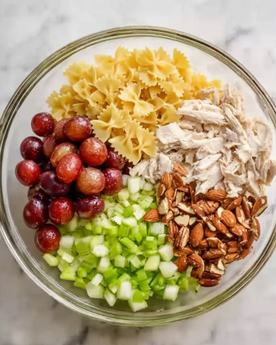 A clear glass bowl filled with six distinct layers arranged next to each other in small sections. Starting at the top is a layer of yellow bowtie pasta with a smooth texture. To the right, shredded white chicken sits in a light pile. Below the chicken, a pile of chopped green celery is visible, with a fresh and crisp look. At the bottom left, there are halved red grapes with shiny skins. Next to the grapes, a layer of rough, brown almond pieces fills the space. The bowl rests on a white marbled surface with natural light illuminating the scene, and a woman's hand slightly touching the bowl's edge. Photo taken with an iphone --ar 4:5 --v 7