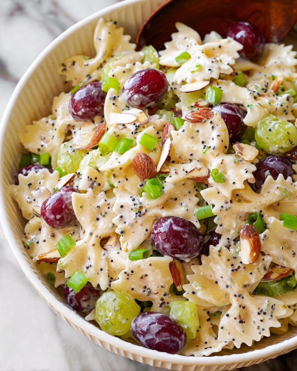 The image shows a close-up of a creamy pasta salad in a white bowl with ridged edges. The salad has light yellow bowtie pasta coated in a creamy white dressing speckled with tiny black poppy seeds. Mixed evenly throughout are dark red and purple grape halves, small green celery pieces, chopped green onions, and sliced almonds adding texture. The ingredients are well combined, creating a colorful and fresh look with the bright creamy sauce coating each layer on a white marbled surface. photo taken with an iphone --ar 4:5 --v 7