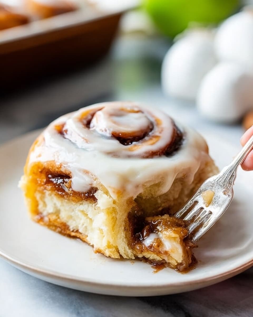 A single cinnamon roll sits in the center of a white plate, showing about three thick layers of soft dough with swirls of brown cinnamon filling inside. The roll is covered with a creamy white icing that softly drips over the edges. At the bottom, a sticky caramel-colored sauce pools and spreads onto the plate, adding a shiny texture beneath the roll. A silver fork with ornate details rests beside the cinnamon roll on the plate. In the background, there are parts of two green apples, one whole and one cut in half, placed on a white marbled surface. photo taken with an iphone --ar 4:5 --v 7