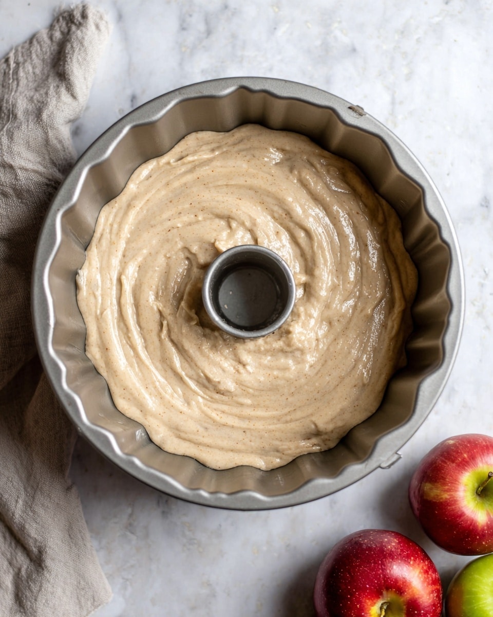 A light brown, thick batter with small specks is spread evenly inside a silver bundt cake pan with scalloped edges and a hollow center, sitting on a white marbled surface. To the bottom right of the pan, three fresh apples with red and green skin add color contrast, and a soft, light gray cloth is partially visible to the left. The batter has a smooth, creamy texture with swirls from spreading, filling the pan almost to the edges. Photo taken with an iphone --ar 4:5 --v 7