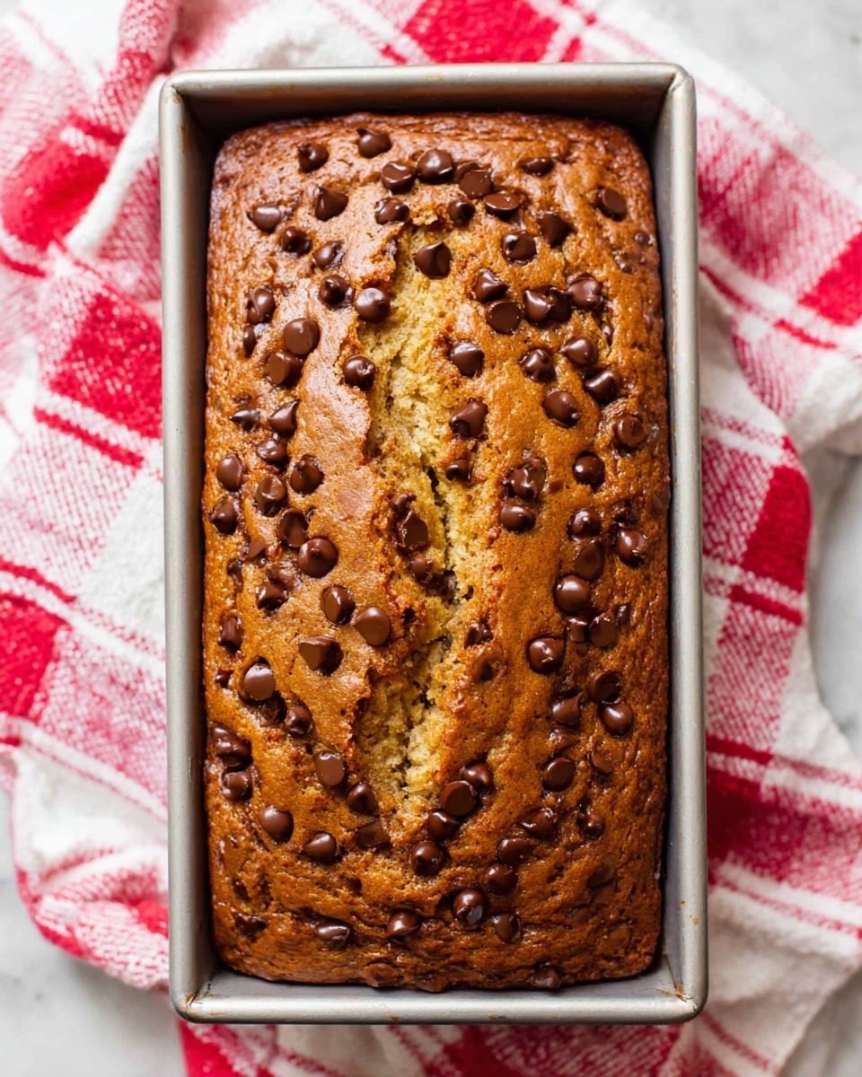 A freshly baked rectangular banana bread loaf with a golden brown crust spotted with many dark brown chocolate chips scattered unevenly on the top. The bread has a crack running down the middle showing a moist light yellow inside. It is in a silver baking pan placed on a white marbled surface with a white and red checkered cloth partially visible underneath. Photo taken with an iphone --ar 4:5 --v 7
