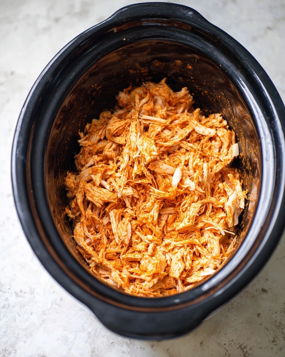 Inside a black slow cooker, there is a single layer of shredded cooked chicken that is orange in color from spices, with some white pieces visible in between. The chicken pieces have a fibrous, tender texture and fill the bottom of the cooker, showing a bit of the smooth, dark inner surface. The slow cooker is placed on a white marbled surface. photo taken with an iphone --ar 4:5 --v 7