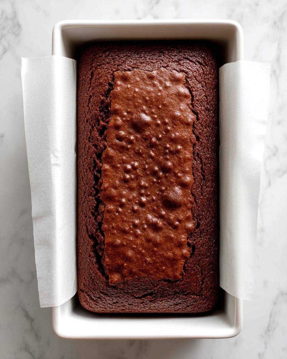 A rectangular chocolate cake with a rich, dark brown color and a slightly bumpy, textured surface sits in a white rectangular baking pan lined with white parchment paper that folds over the edges. The cake fills most of the pan and looks firm and moist. The background is a white marbled texture. photo taken with an iphone --ar 4:5 --v 7