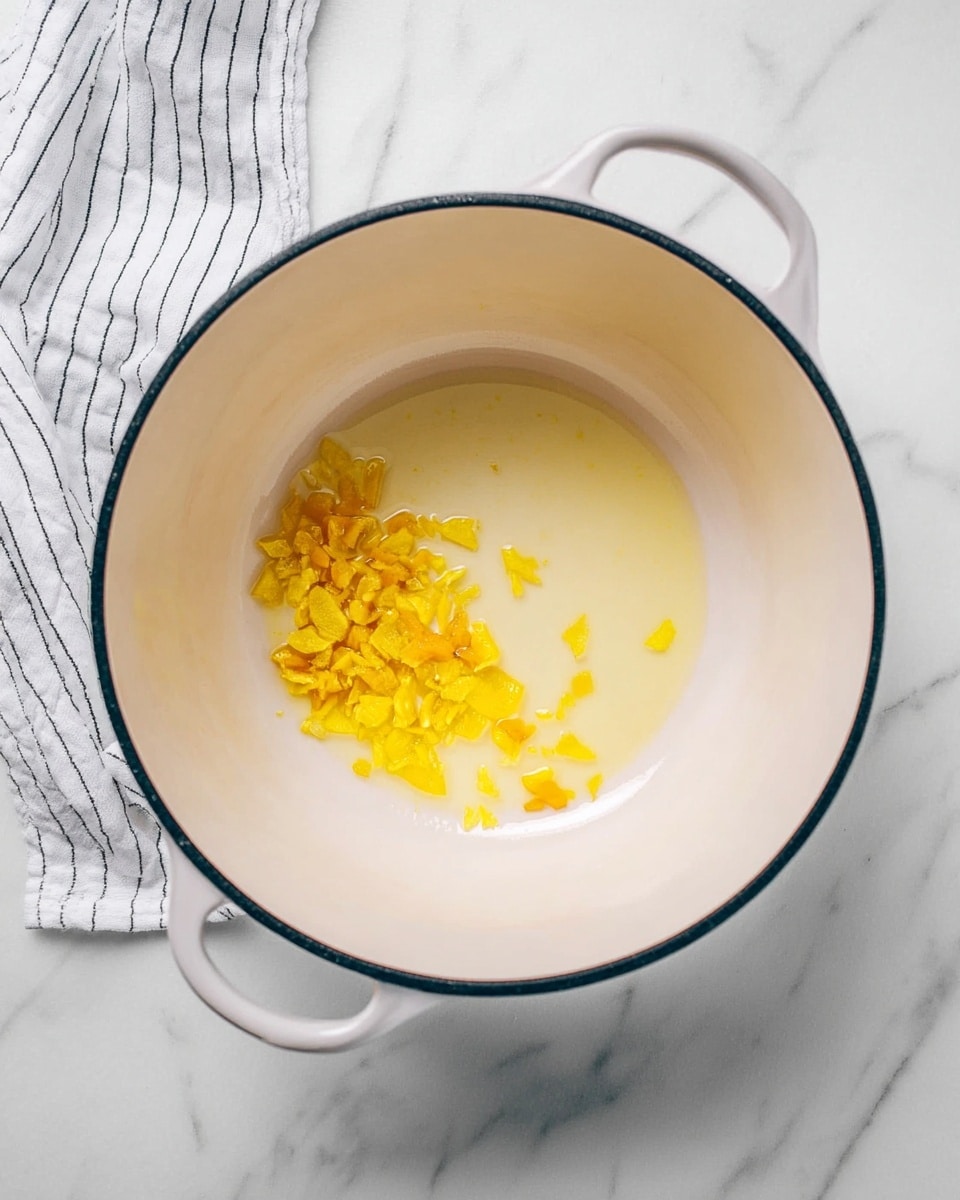 Inside a white pot with a dark rim, small yellow pieces of a vegetable or fruit sit on the right side in melted yellow butter. The pot is placed on a white marbled surface with a white and black striped cloth partly visible near the handle. The colors are soft, focusing on the light yellow inside the pot against the clean white background. photo taken with an iphone --ar 4:5 --v 7