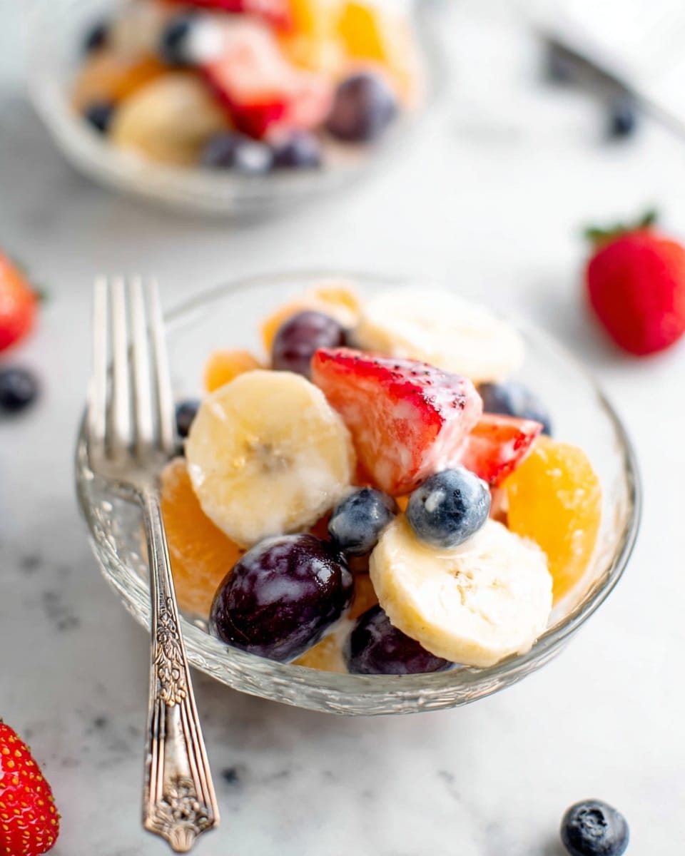 A small pile of mixed fruit salad sits in a clear glass bowl on a white marbled surface, featuring layers of orange segments, round slices of pale yellow banana, red strawberry slices, deep blue blueberries, and dark purple grapes cut in half, all lightly coated with a creamy white dressing that adds a glossy look. A silver fork with ornate details rests on the left side of the bowl. Scattered around the bowl are whole strawberries and blueberries in soft focus. Another similar bowl is blurred in the background. Photo taken with an iphone --ar 4:5 --v 7