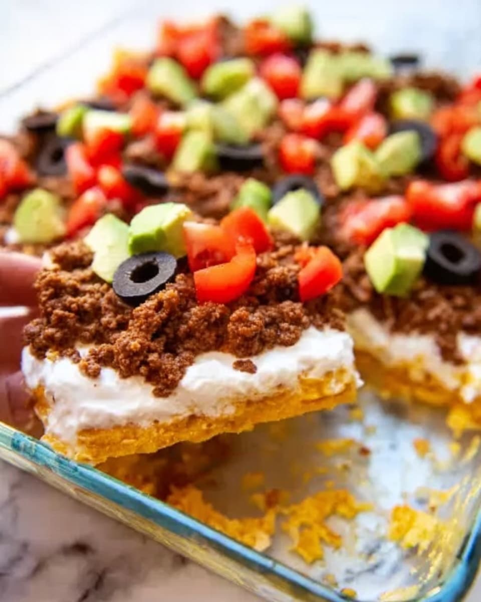 The dish appears to be a layered dip served in a clear glass rectangular dish placed on a white marbled surface. The bottom layer is a golden-yellow base, likely a chip or tortilla layer, topped by a thick smooth white layer that looks like sour cream or cream cheese spread evenly. Above this, there is a generous layer of cooked ground meat in brown color with a crumbly texture, spread across the dish. Scattered over the meat layer are small cubes of bright green avocado and chunks of red tomato, along with sliced black olives for contrast. A woman's hand is seen holding a piece of the dish on the left side. Photo taken with an iphone --ar 4:5 --v 7