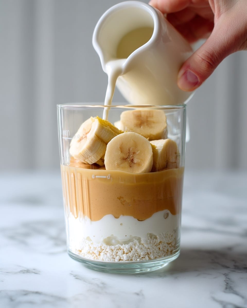 The image shows a transparent glass measuring cup placed on a white marbled surface. Inside the cup, there are three layers: the bottom layer is thick and white with a rough texture, resembling cottage cheese or yogurt; the middle layer is smooth and light brown, looking like peanut butter; and the top layer contains several thick pieces of pale yellow banana stacked loosely. Above the banana pieces, a woman's hand is pouring a light cream or milk from a white jug into the cup. The background is soft and light, keeping the focus on the measuring cup and its contents. photo taken with an iphone --ar 4:5 --v 7