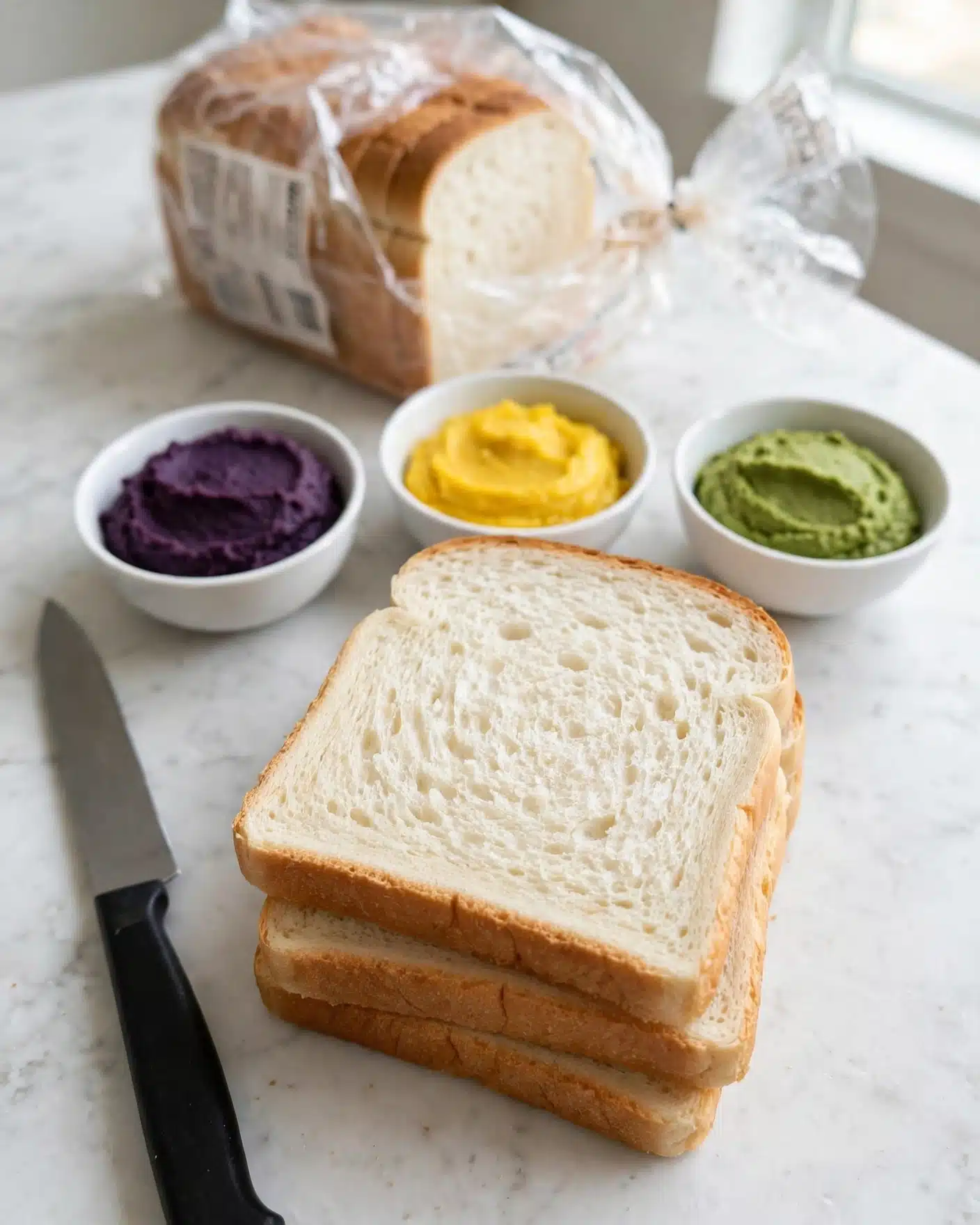 Rainbow Ribbon Sandwiches with Colorful Vegetable Fillings Recipe 4 The image shows a stack of three plain white sandwich bread slices with soft texture and light brown crust at the edges, placed on a white marbled surface. Behind the bread, there are three white bowls filled with colorful pastes: a deep purple paste on the left, a bright yellow paste in the middle, and a green paste on the right. A partially opened bread bag with more slices is visible in the background, and a knife with a black handle is placed on the left side of the bread stack. Photo taken with an iphone --ar 4:5 --v 7