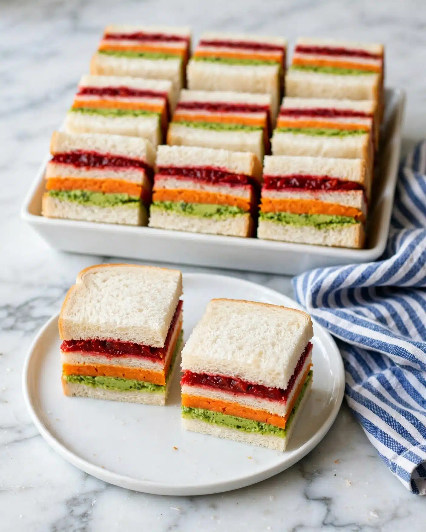 Rainbow Ribbon Sandwiches with Colorful Vegetable Fillings Recipe 5 The image shows small square sandwiches stacked with three layers of white bread. Between the bread layers are three colorful fillings: bright green, vivid orange, and deep red, arranged in horizontal stripes. Two of these sandwiches are placed on a white round plate in the front, while behind them there is a white rectangular dish filled with more stacked sandwiches. A blue and white striped cloth is casually placed near the plate, and the scene is set on a white marbled surface. Photo taken with an iphone --ar 4:5 --v 7