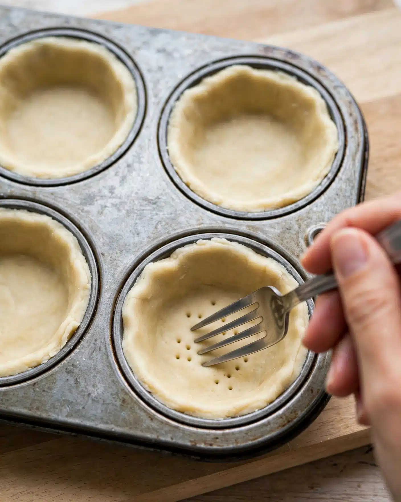 A close-up view of a metal muffin tin with four visible small tart shells made from pale dough pressed into each cup. A woman's hand is holding a fork, poking holes into the dough inside one of the shells to prepare it for baking. The muffin tin is resting on a wooden cutting board with a light finish, visible in the background. The dough has a soft, smooth texture with a slightly uneven edge. photo taken with an iphone --ar 4:5 --v 7