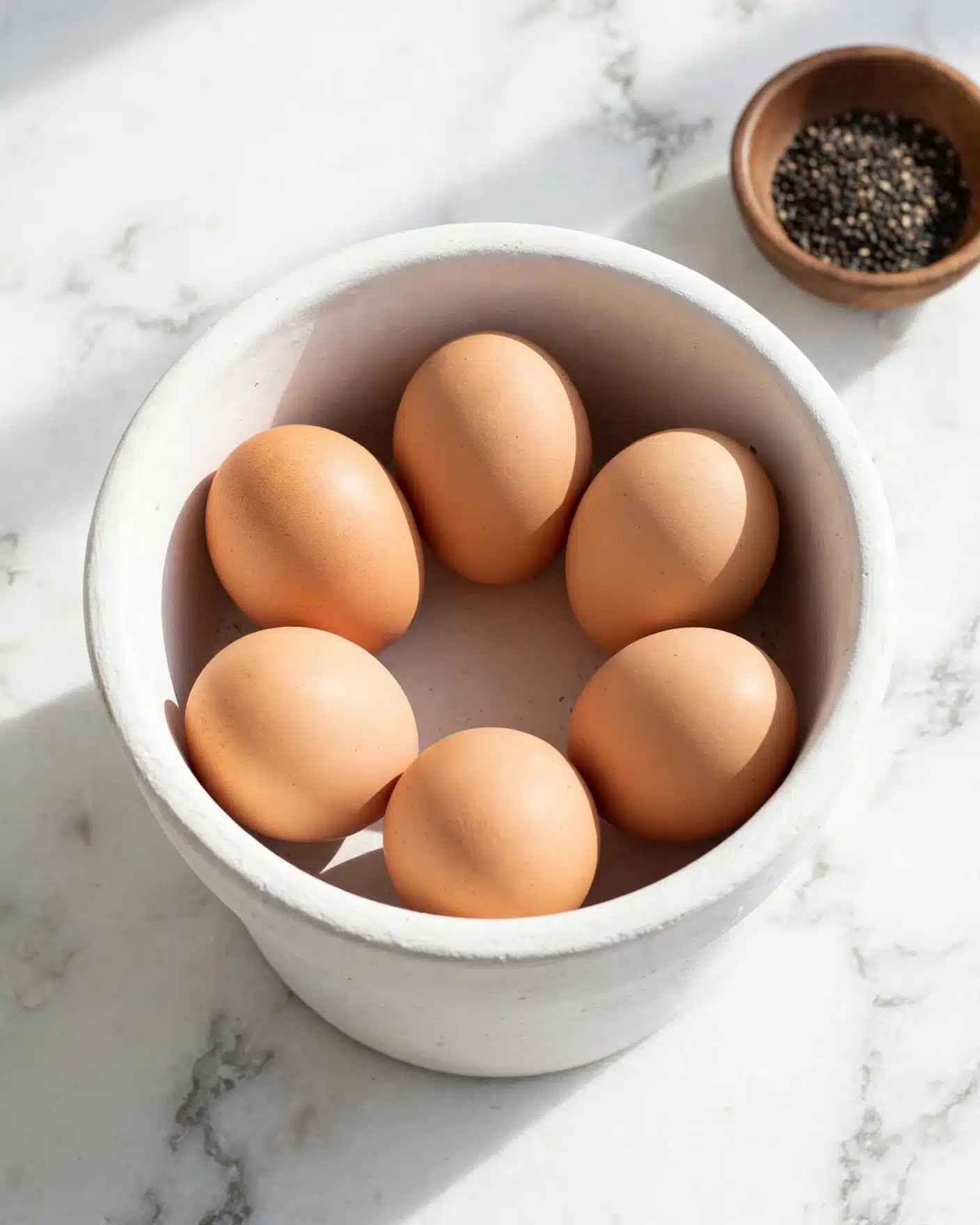 A white pot filled with six light brown eggs resting on a white marbled surface. The eggs are arranged loosely in a circular shape inside the pot. In the upper right corner, there is a small brown bowl with black pepper seeds. The overall lighting is bright and natural, emphasizing the smooth texture of the eggs and the clean white pot. Photo taken with an iphone --ar 4:5 --v 7